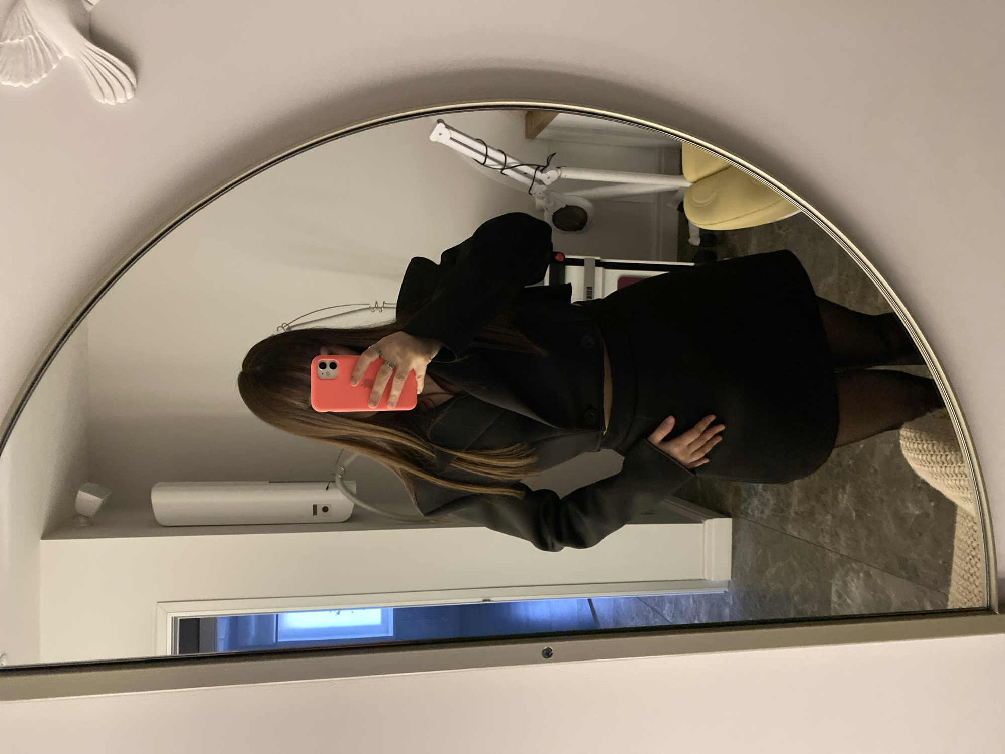 A mirror selfie taken in an indoor setting in Votkinsk, Russia, depicts a person, likely female, obscuring her face with a coral-pink smartphone. She has long brown hair and is dressed in a black long-sleeved jacket or blazer, a black skirt, and black tights. Her left hand holds the phone, revealing a ring on one finger, while her right hand rests on her hip.
The image is captured through an arched mirror, reflecting parts of the room. To the upper left of the mirror, a white decorative wall accent, resembling a bird or wing, is visible. Inside the mirror's reflection, behind the person, a white device, possibly an air freshener or humidifier, rests on a shelf. Below this, a section of a door or hallway with a bluish light is reflected. A partial view of a yellow armchair or seat is seen towards the upper right within the reflection. The floor has a grey, marbled texture, and a beige, textured item, possibly a rug or blanket, is visible in the lower right corner of the reflection. The lighting suggests an indoor setting, possibly using artificial light. There is no visible text in the image.
