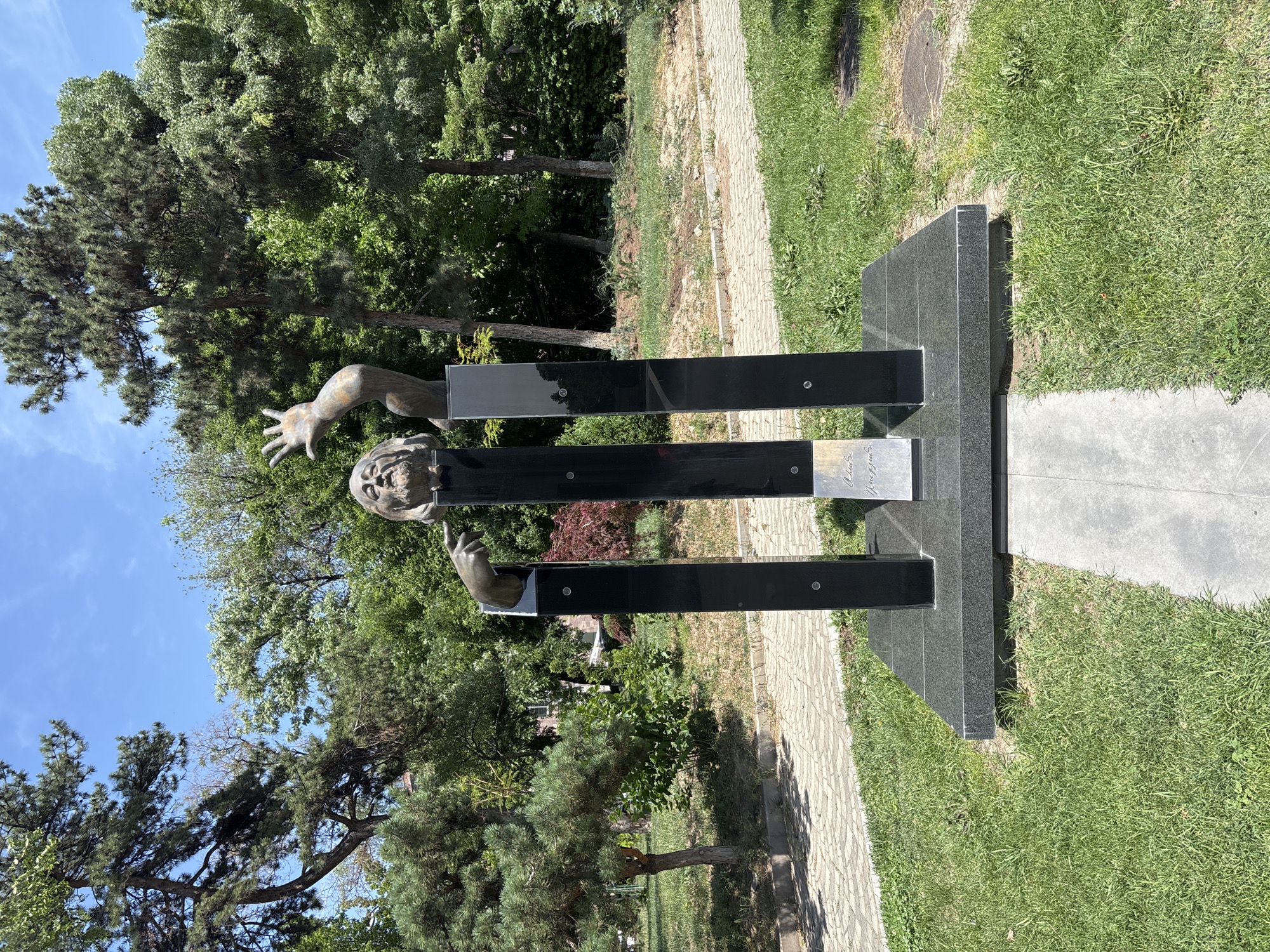 The image shows a sculpture in Yerevan, Armenia, located in the vicinity of the Cascade complex. The sculpture is made of bronze and granite and depicts a man's head and torso emerging from three black granite pillars. The man has his eyes closed, and his mouth is slightly open. One hand is raised in the air, palm open, while the other hand points to the side. The pillars are arranged in a triangular formation, with the man's head at the apex. The sculpture is set on a granite base, surrounded by a paved pathway and lush green grass. Trees and foliage form the background. The sky is clear and blue, indicating a sunny day.