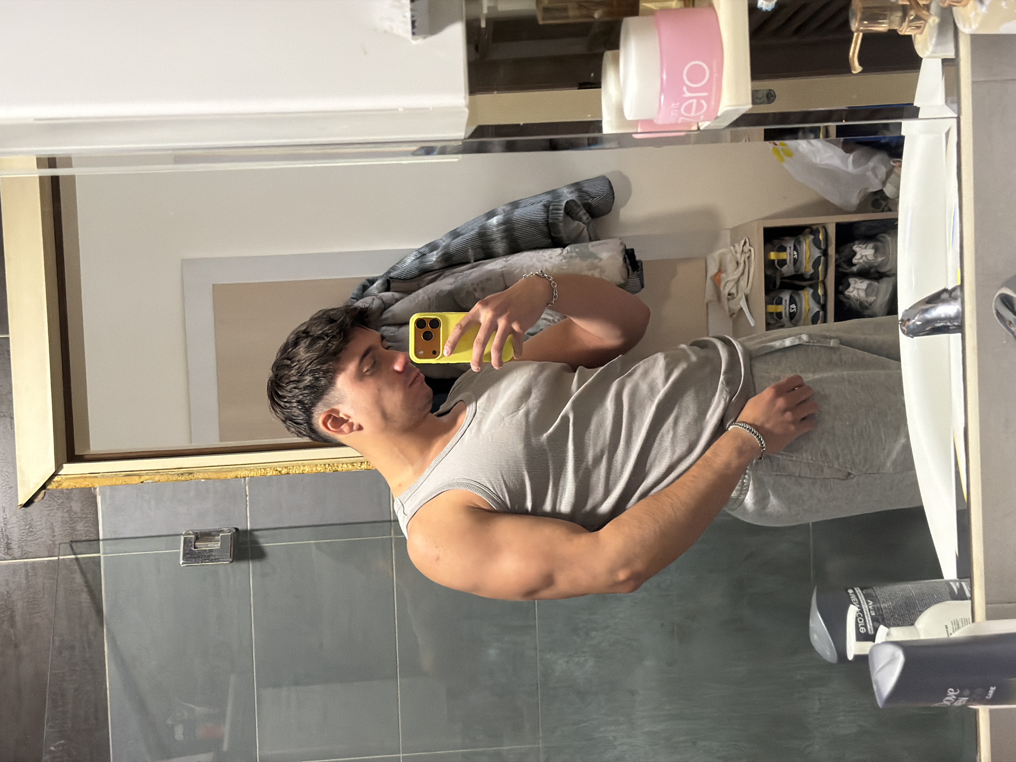 A man is taking a selfie in a bathroom mirror. He is wearing a grey tank top and grey sweatpants. The bathroom has grey tile walls. There is a white sink and various toiletries visible in the mirror. The city of Sofia, Bulgaria is not discernible from the image.