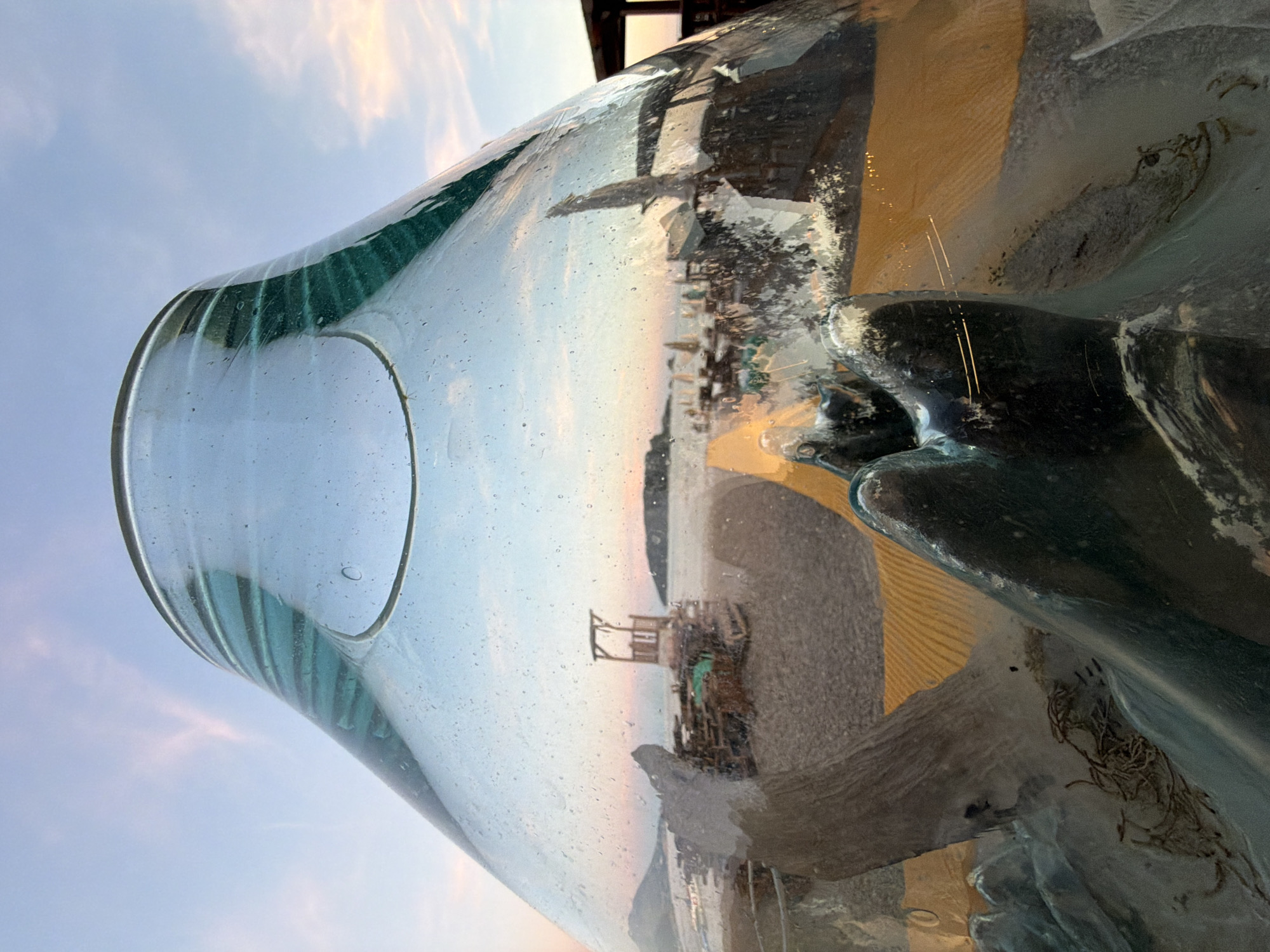 The image is a close-up, low-angle shot of a large, transparent glass object that distorts the reflection of its surroundings. The glass object is placed on a beach, likely in Budva, Montenegro, given the presence of a lifeguard tower and beach umbrellas visible in the reflection. The sky is a pale blue with soft pink clouds, indicating either sunrise or sunset. The beach has a sandy surface, and there are wooden structures and beach furniture. The glass itself has a thick, uneven texture with some bubbles and imperfections, and it curves inwards at the top, creating a funnel-like opening. The reflection shows the beach, the sea, and the sky, warped and fragmented by the glass. In the foreground, within the glass, are dark, amorphous shapes that appear to be part of the glass object's base or internal structure. Some strands of seaweed are visible on the sand near these shapes. The overall impression is one of a surreal and artistic interpretation of a beach scene, captured through a unique glass lens.