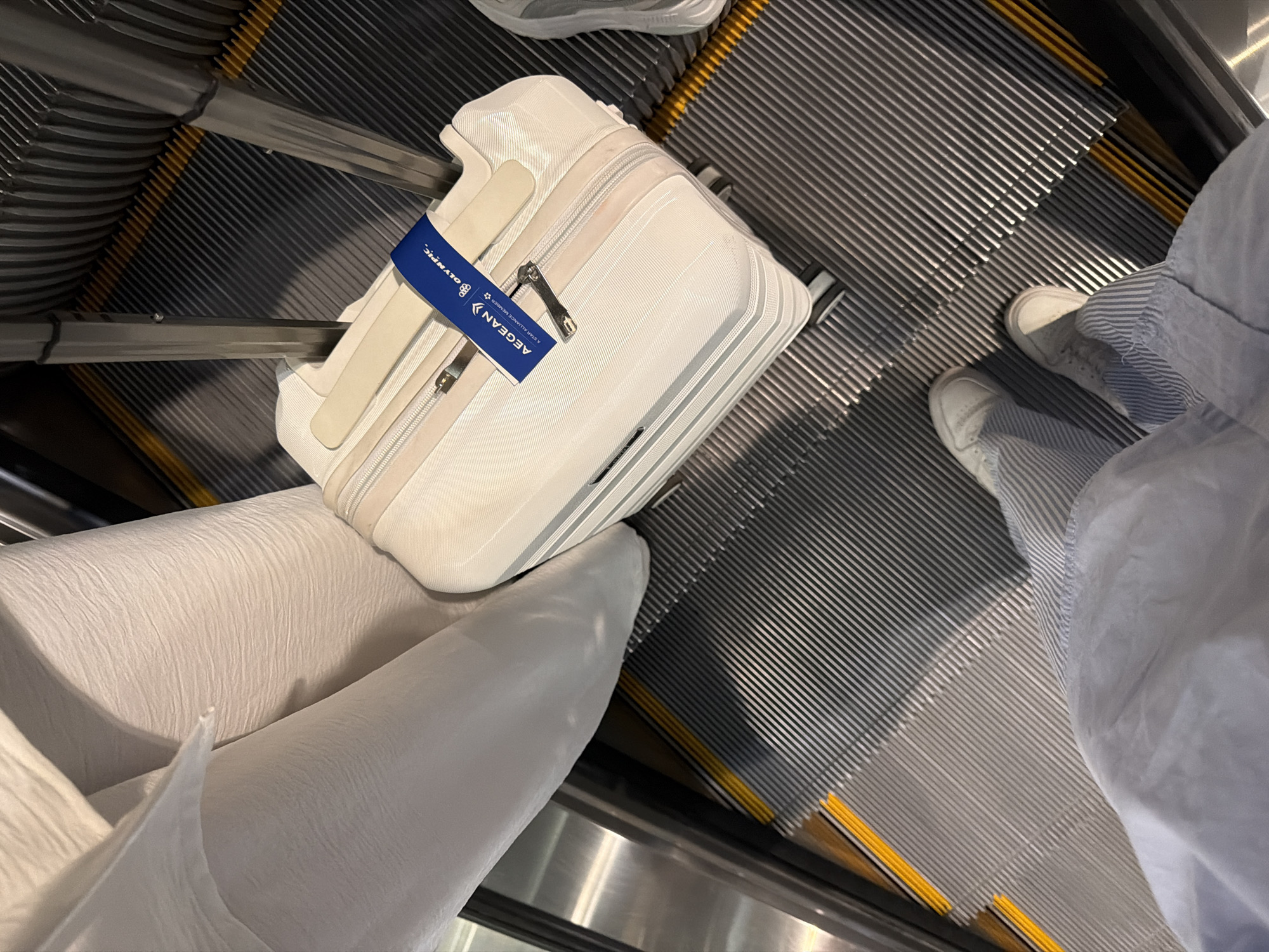 The image captures a scene on an escalator, featuring a person with a white suitcase. The suitcase is labeled with a blue tag bearing the words "AEGEAN Olympic, A STAR ALLIANCE MEMBER." The person is wearing light-colored sneakers and a light blue patterned shirt. The escalator has metal steps with yellow accents. The location is not discernible from the image.