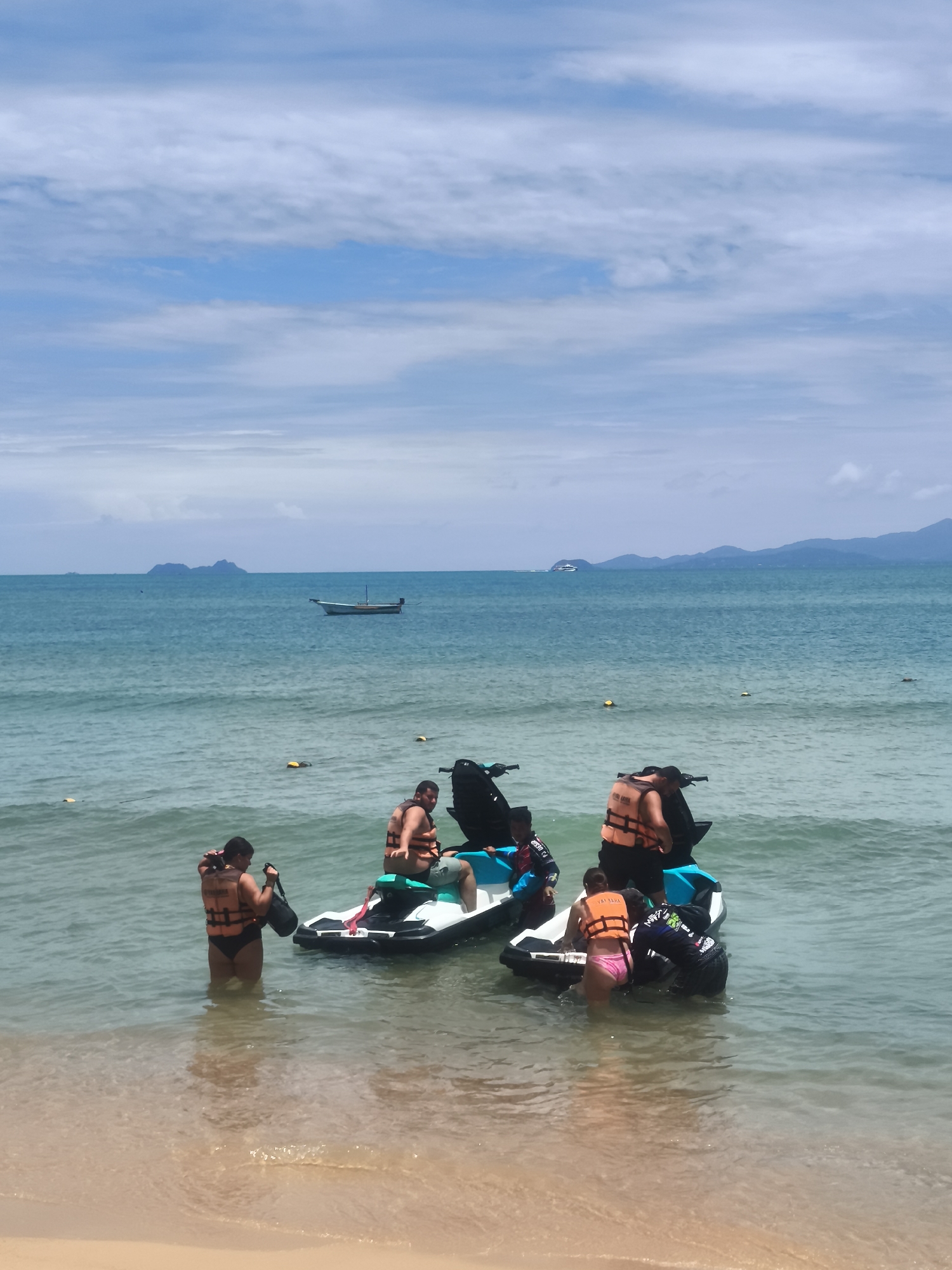 The image depicts a beach scene in Ko Samui District, Thailand, under a partially cloudy sky. Several people are interacting with jet skis in the shallow turquoise water. On the left, a person wearing an orange life vest and black bottoms stands in the water, holding a black bag. Next to them, a man in an orange life vest is seated on a jet ski. To the right, another jet ski has two people on it, one in an orange life vest and another with a child in a blue and orange life vest. In the foreground, the sandy beach meets the water. In the middle ground, a small white boat with a wooden hull is anchored in the distance, with islands visible on the horizon. Yellow buoys are scattered in the water.