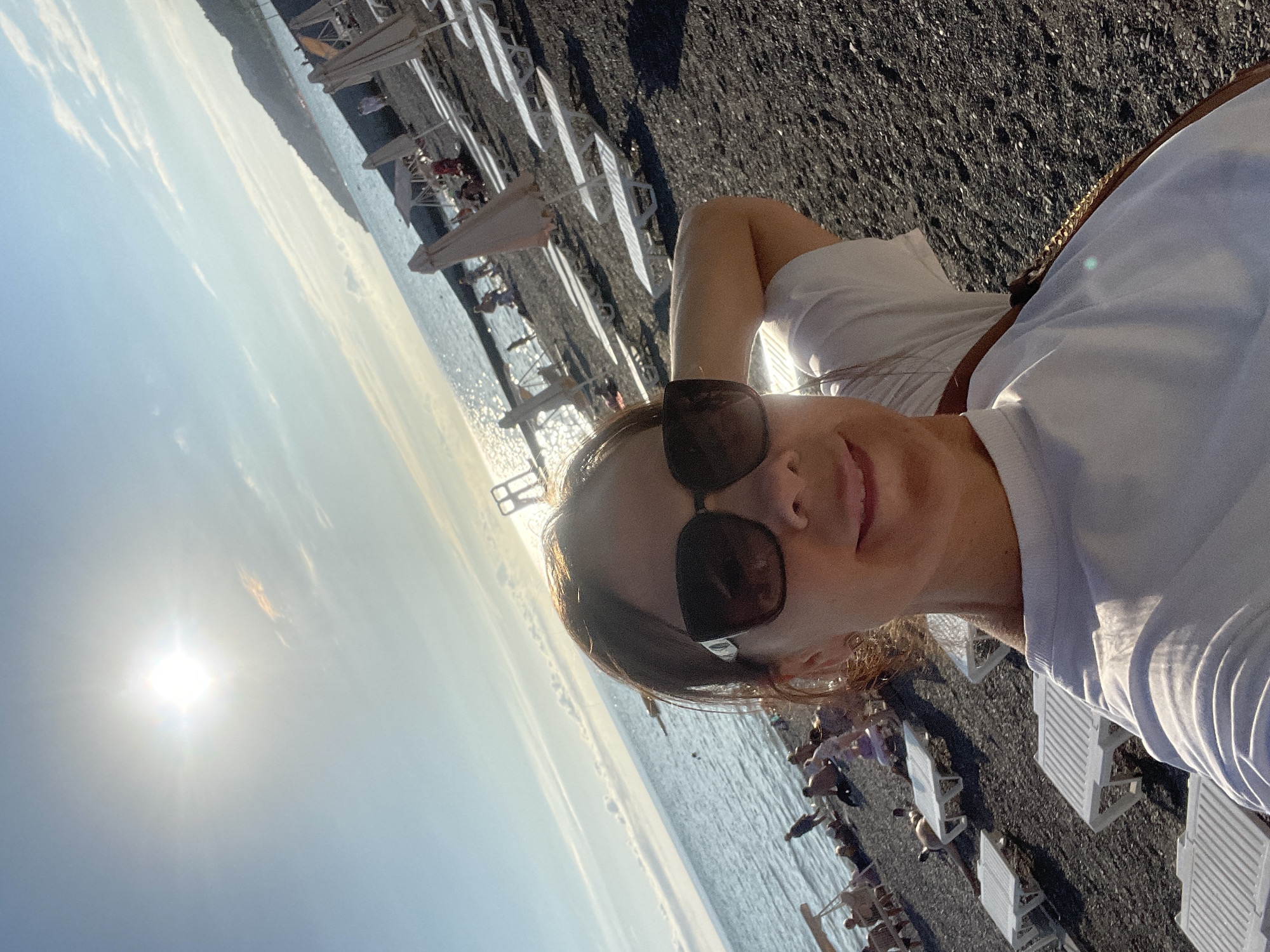 A woman in sunglasses and a white t-shirt takes a selfie on a pebble beach in Sochi, Russia. The sun is bright in the sky, and the sea is to her left. Many empty lounge chairs are visible on the beach, along with a few people in the distance. The background features a coastline with hills.