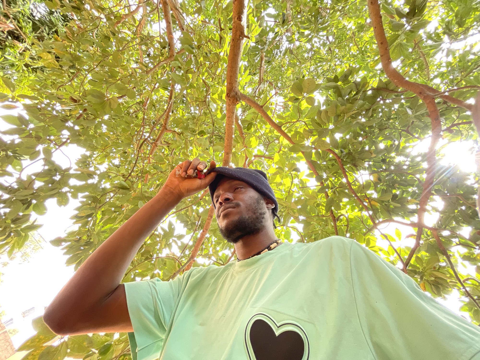 A low-angle shot depicts a man wearing a black beanie, a light green t-shirt with a black heart on it, and a beaded necklace standing under a tree with dense foliage. He is holding the brim of his hat with his right hand. The location appears to be outdoors, likely in a park or green space. The background is filled with tree branches and leaves, with sunlight filtering through them. While Jos, Nigeria, is mentioned, there is no visible indicator in the image to confirm the location with certainty, such as landmarks or local signage.