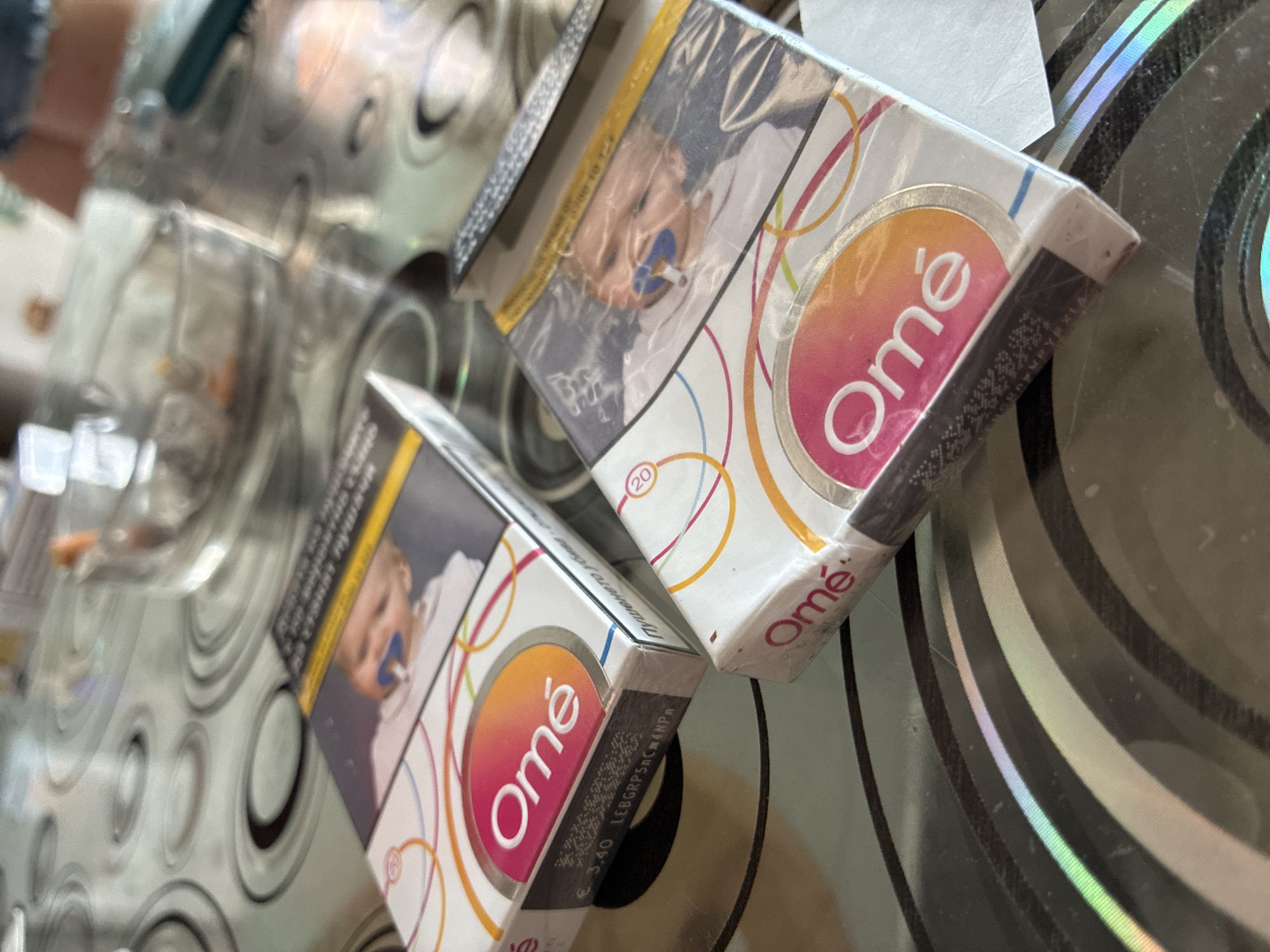 The image shows two packs of "Omé" brand cigarettes. The packs feature images of a baby with a pacifier on the front and health warnings on the side. They are placed on a reflective surface, possibly a glass table with a patterned design. Based on the brand of cigarettes, it's possible the image was taken in Varna, Bulgaria. There are some reflections of other objects in the background, but they are indistinct.