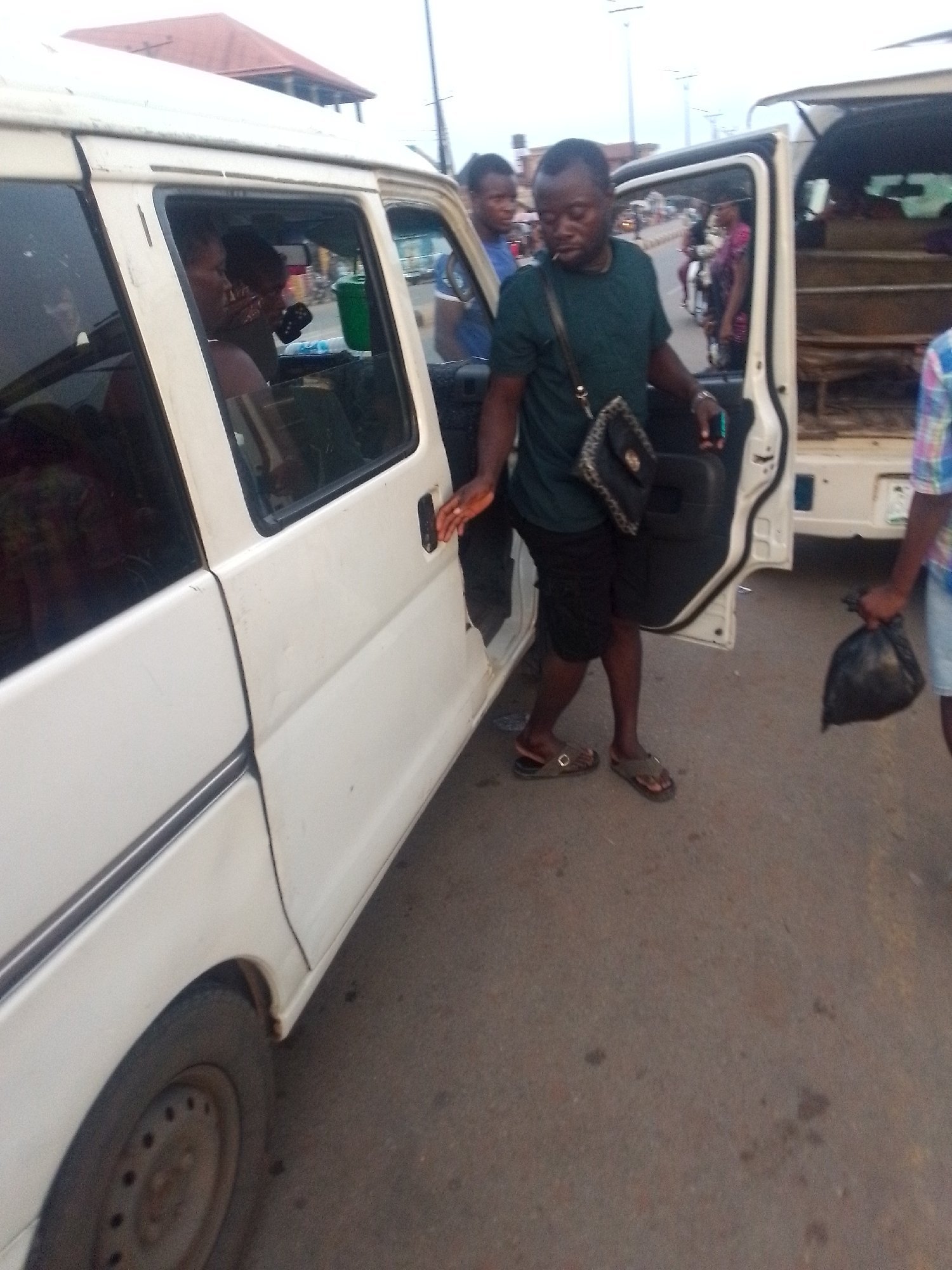 A man in a dark green shirt, black shorts, and brown sandals is exiting a white van in a city in Unknown, Unknown. He is holding the door with his left hand and has a black crossbody bag on his left shoulder. Another man stands nearby holding a black plastic bag. People can be seen inside the van and in the background on the street. Other vehicles, including a motorcycle, are visible in the background.