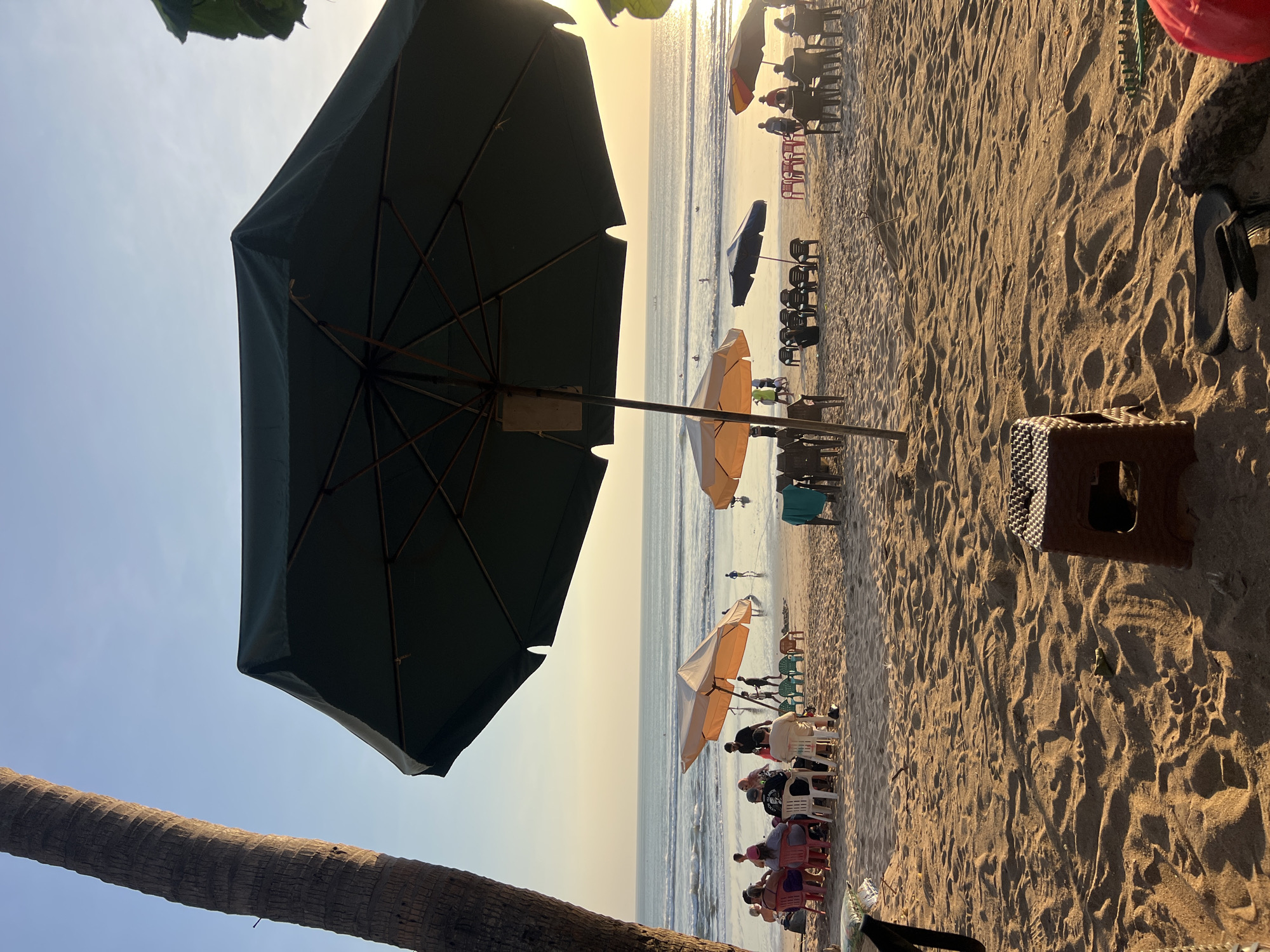 The image depicts a beach scene at sunset in Kuta, Indonesia. The foreground is dominated by sand and a small brown plastic stool. In the mid-ground, several groups of people are seated at tables under beach umbrellas, suggesting a relaxed dining or socializing atmosphere. The umbrellas vary in color and size, with a large dark green one prominently featured in the foreground, partially obscuring the view. Beyond the seated patrons, the ocean stretches to the horizon, with gentle waves breaking on the shore. A few individuals can be seen wading or swimming in the water. The sky is a soft gradient of blue and orange, indicating the approaching sunset, and a palm tree trunk is visible on the left side of the frame, reinforcing the tropical location. The overall impression is one of a tranquil evening by the sea, with a focus on outdoor leisure activities.