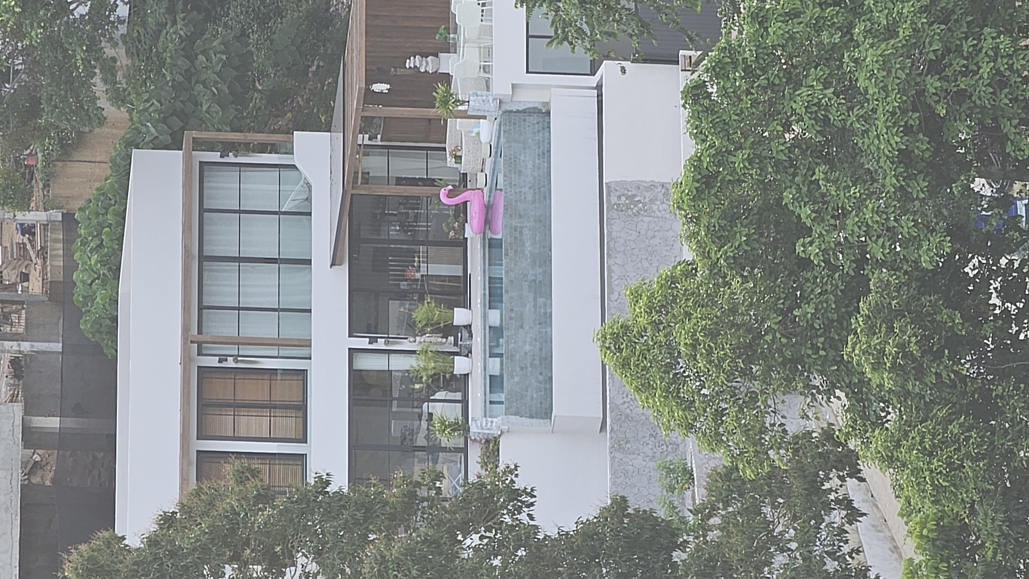 A pink inflatable flamingo floats in a swimming pool on the rooftop of a multi-story modern building. The building has large windows and is surrounded by lush green trees. In the background, construction is visible on a hillside. The scene appears to be a resort or private residence in Baan Chaweng, Thailand, a popular tourist area known for its beaches and nightlife. The pool area is furnished with white outdoor seating.