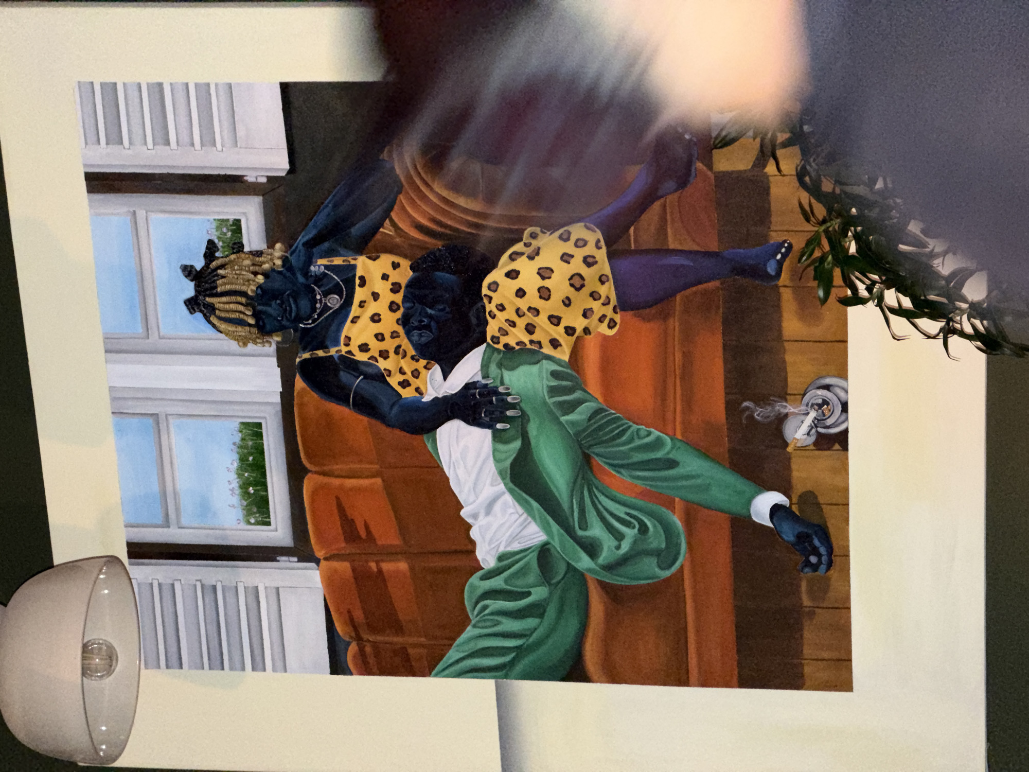 A painting depicting two figures is framed. The figure on the left wears a yellow leopard-print dress and has blonde dreadlocks. The figure on the right is wearing a green suit with a white shirt underneath. They appear to be in an embrace. In the background, there is a window with a white frame and a view of a blue sky and green foliage. To the right, a cigarette burns in an ashtray atop a wooden surface. The setting resembles an interior with an orange-colored sofa in the background. A spotlight illuminates the left side of the painting. The image was taken in Durbanville, South Africa.