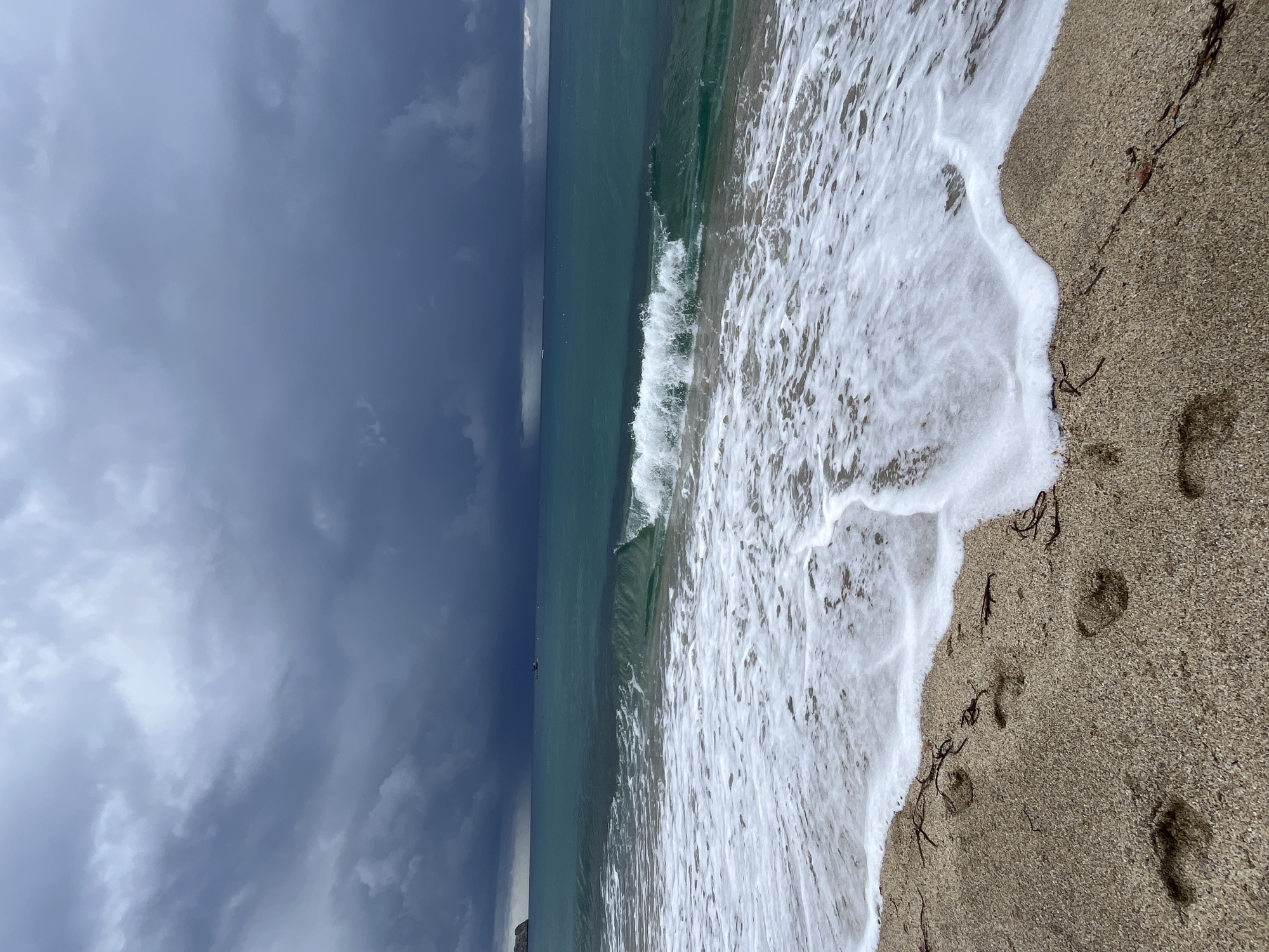 The image depicts a beach scene in Alanya, Turkey. A sandy shoreline is visible on the right side of the frame, marked with a few footprints and scattered bits of seaweed. The tide is coming in, with white foamy waves washing onto the sand. The water in the middle of the frame is a murky green-blue, with a single wave breaking. On the left side, a dark and cloudy sky suggests an approaching storm.