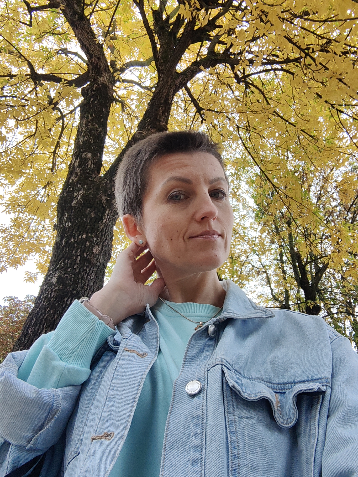 A woman with short, grey hair and blue eyes is looking at the camera, with her left hand touching her neck. She is wearing a light blue denim jacket over a teal sweatshirt. Behind her, a large tree with yellow leaves is visible, suggesting an autumn setting. The scene appears to be outdoors, possibly in a park or residential area within Minsk, Belarus.