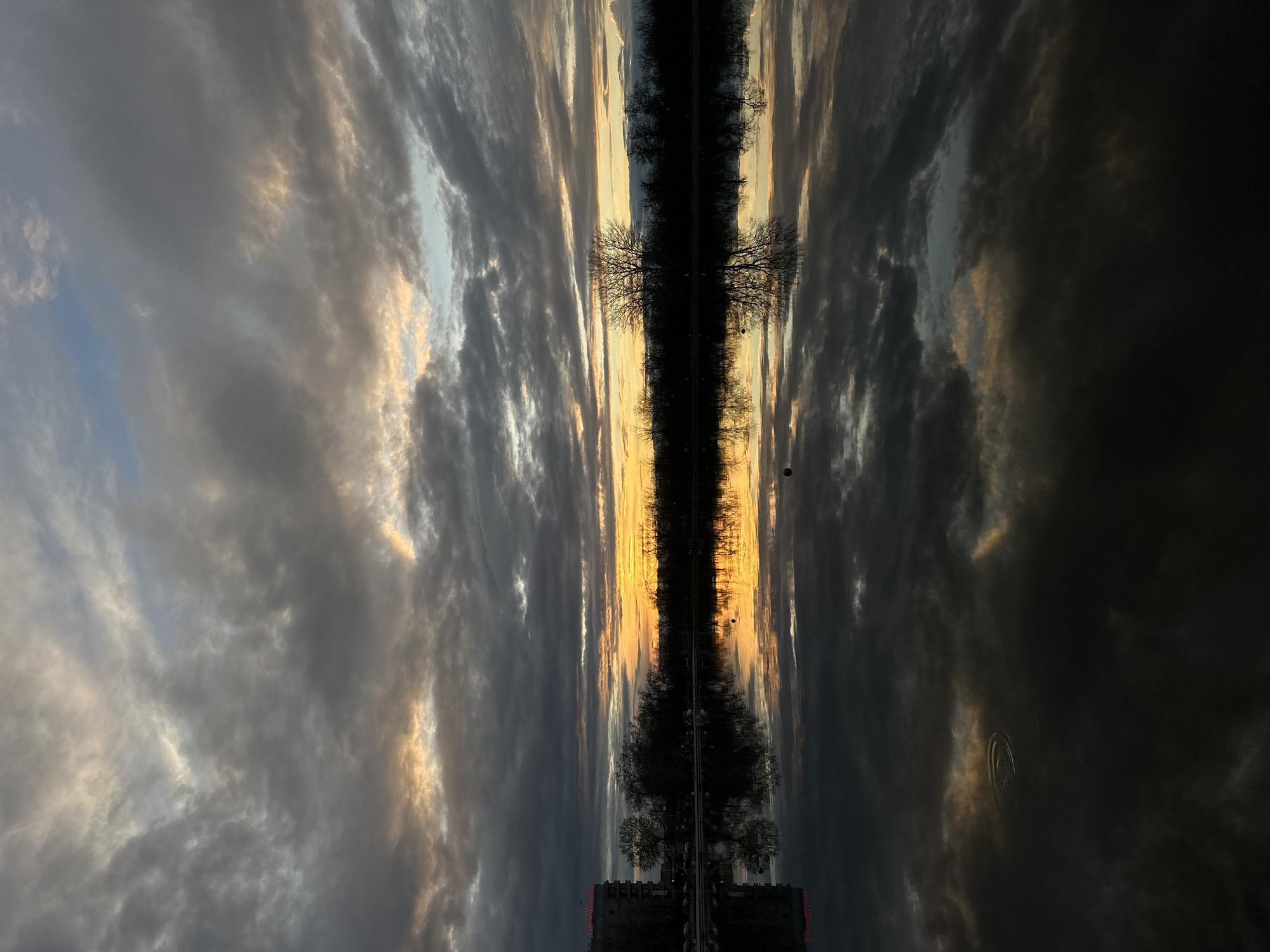 The image shows a scene in Plovdiv, Bulgaria, reflected in a body of water. The sky above is filled with dark, textured clouds, partially illuminated by the colors of a sunset. Trees line the central section, their bare branches silhouetted against the sky. A building with red lights is visible at the bottom. There are also some ripples on the surface of the water.