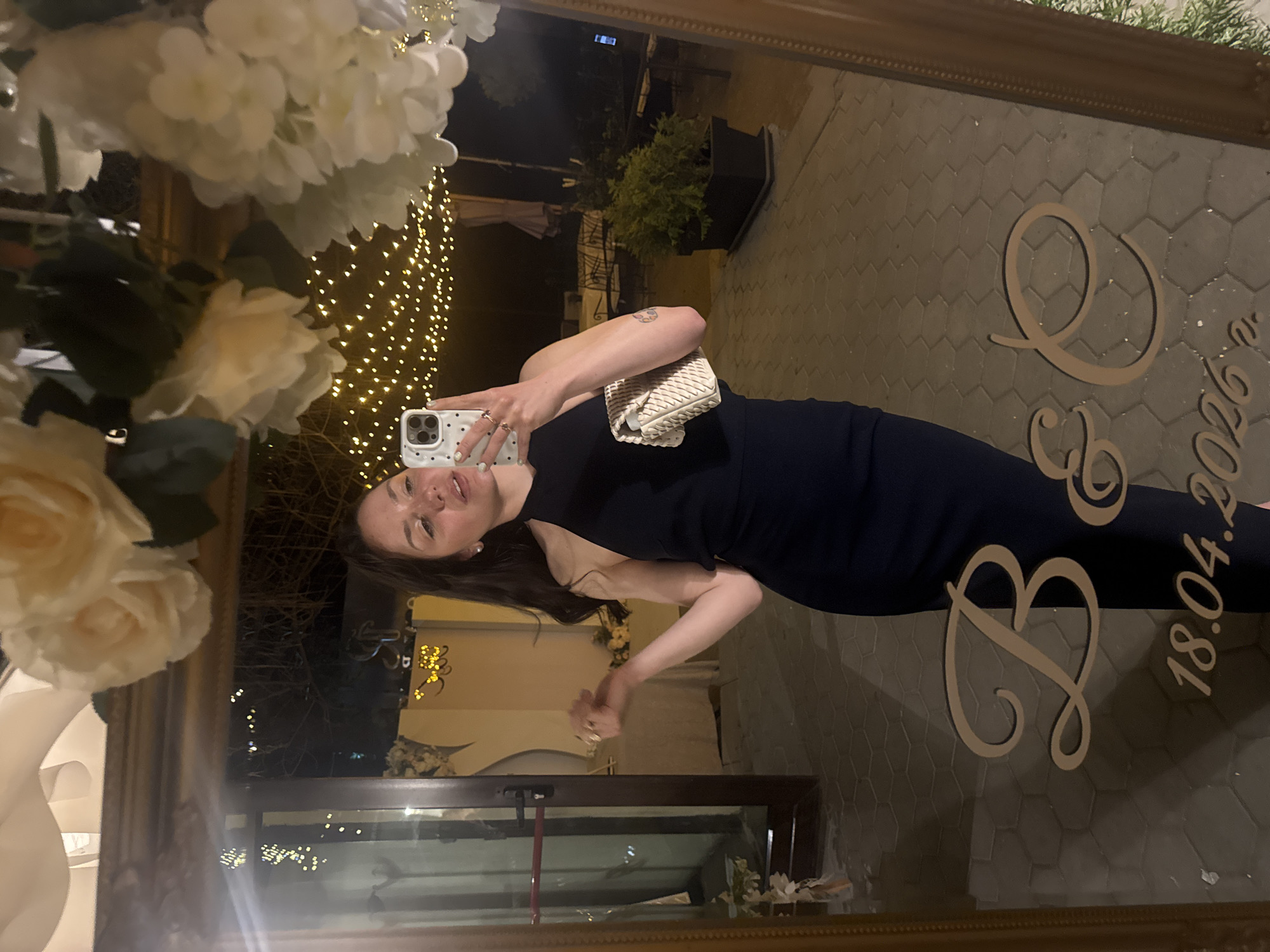 A woman takes a selfie in front of a mirror that reflects an outdoor event space in Blagoevgrad, Bulgaria. The mirror reflects her dark sleeveless dress, fair skin, and a small purse. The reflection behind her shows outdoor lighting, greenery, and a sign with "B & C 18.04.2026." A floral arrangement is in front of the mirror.