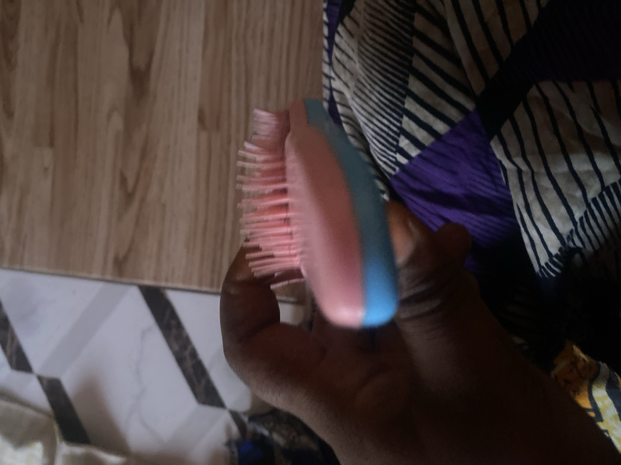 A hand is holding a pink and blue hairbrush against a background of patterned fabric and wooden flooring. The hairbrush is small and oval-shaped with pink bristles. The hand is dark-skinned and appears to be in focus. The fabric in the background has a striped pattern with black, white, and purple sections. The wooden flooring is light brown with visible grain. The scene is indoors, possibly in a residential setting. There is no identifiable information about Jauro Yinu, Nigeria in the image.