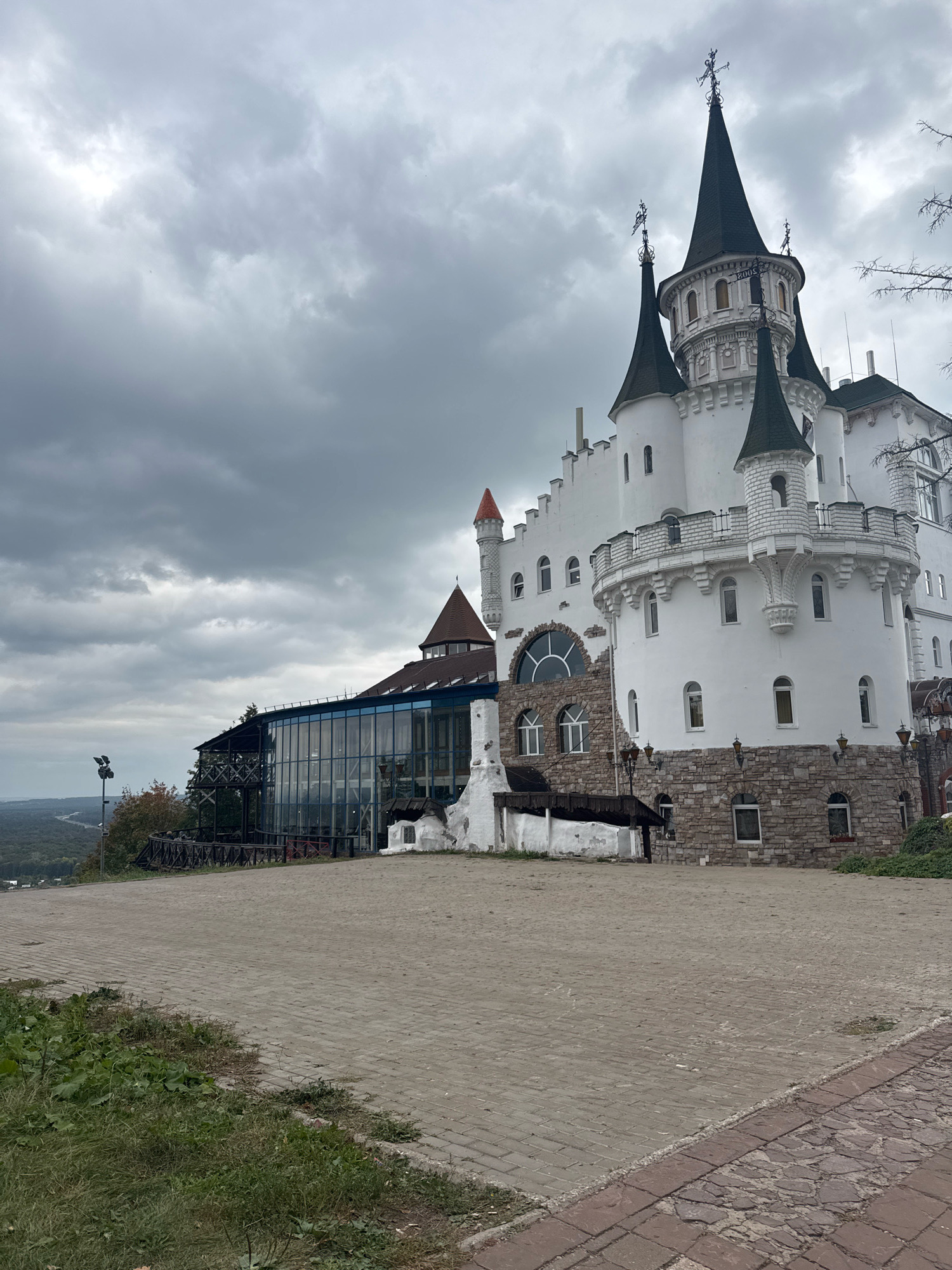 This is an outdoor, eye-level, medium shot of a white, fairy-tale-style castle. The castle has multiple pointed turrets with dark green roofs. The building is made of white stone and has many windows of varying sizes. At the base of the castle, there is a section made of rough-hewn stone blocks, with arched windows. To the left of the castle, there is a modern glass-enclosed building with a dark roof, possibly a restaurant or event space, and a wooden balcony. The castle is situated on a hill overlooking a forested area with a river or road visible in the distance. The sky is overcast with heavy, grey clouds. The foreground is a paved area made of light grey bricks, with a border of darker, reddish-brown bricks and some patches of grass. There are no people visible in the image. This castle is known as the "Fairy Tale Castle" and is located in the city of Ufa, Russia.