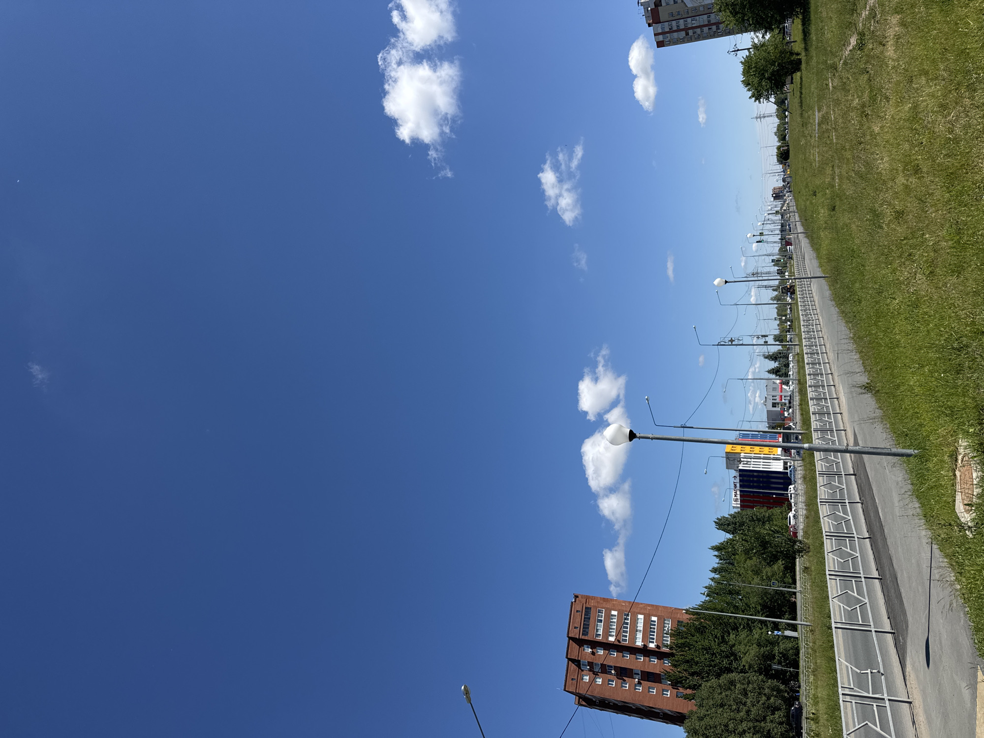 This media file captures a wide-angle view of a sunny urban or suburban landscape. The image is dominated by a bright blue sky, scattered with fluffy white cumulus clouds, occupying roughly the top half of the frame.
In the bottom left, a tall, reddish-brown multi-story building with numerous white-framed windows stands prominently, partially obscured by green trees. To its right, a paved sidewalk or service road stretches into the distance, lined on one side by a white metal fence with a diamond pattern. Beyond this fence is a strip of vibrant green grass.
Along the right side of the road, multiple streetlights with globe-shaped luminaires extend linearly, suggesting a long thoroughfare. Further in the background, a wider road or highway with vehicle traffic is visible, bordered by more trees and additional buildings. Some of these distant buildings are brightly colored, featuring yellow, blue, and red sections. On one of these colorful buildings, the Cyrillic text "СТАЛИТ" (STALIT) is faintly visible.
The scene depicts a clear, bright day, likely midday, with strong sunlight casting distinct shadows. There are no people visible, and no specific activity is taking place, creating a sense of quiet urban expanse. The overall impression is one of a typical, developed city or town area on a pleasant day.