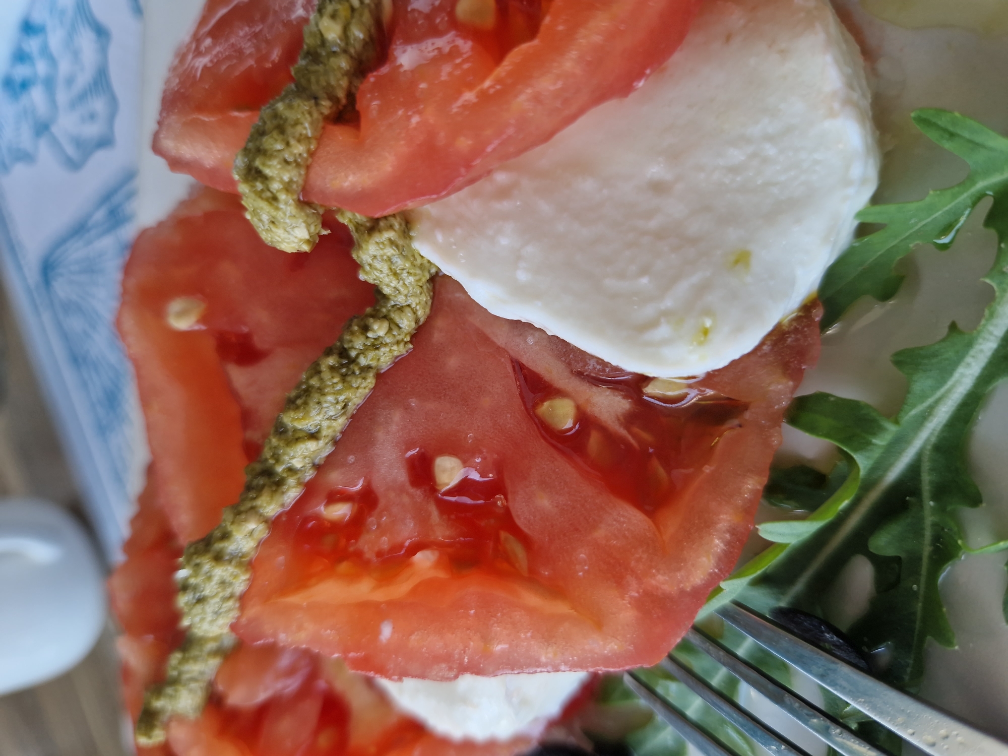 This is a close-up, eye-level shot of a Caprese salad presented on a white plate. The salad features thin slices of ripe, red tomatoes alternated with thick slices of fresh, white mozzarella cheese. A dollop of vibrant green pesto is drizzled over the top of the tomatoes. The salad is arranged on a bed of arugula, with hints of a fork and a black olive visible on the left side of the frame. In the blurred background, a white ceramic cup and a blue and white patterned placemat suggest a dining setting. The image appears to have been taken in a restaurant or cafe in Pomorie, Bulgaria, a city known for its coastal scenery and local cuisine.