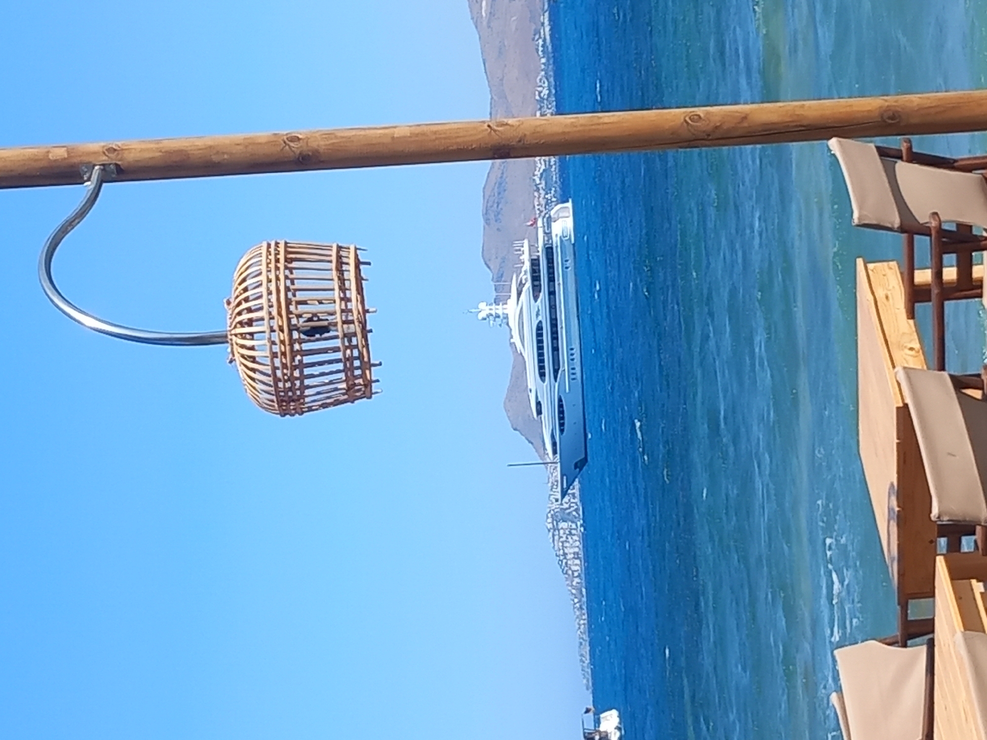 The image depicts a serene coastal scene under a clear blue sky. In the foreground, two wooden tables and chairs are set up, suggesting an outdoor dining area. A prominent wooden pole stands to the right, supporting a decorative, cage-like lantern made of woven material, which hangs from a curved metal arm.
Beyond the seating area, the deep blue sea stretches out, with gentle waves creating a textured surface. In the middle ground, a large white yacht is anchored or cruising in the water. The yacht is sleek and modern, with multiple decks and windows.
In the background, a coastal town with white buildings is nestled against rolling hills. The landscape is characteristic of islands in the Aegean Sea, like Kos, Greece. The overall impression is one of a tranquil, idyllic seaside location, likely a restaurant or cafe overlooking the water.