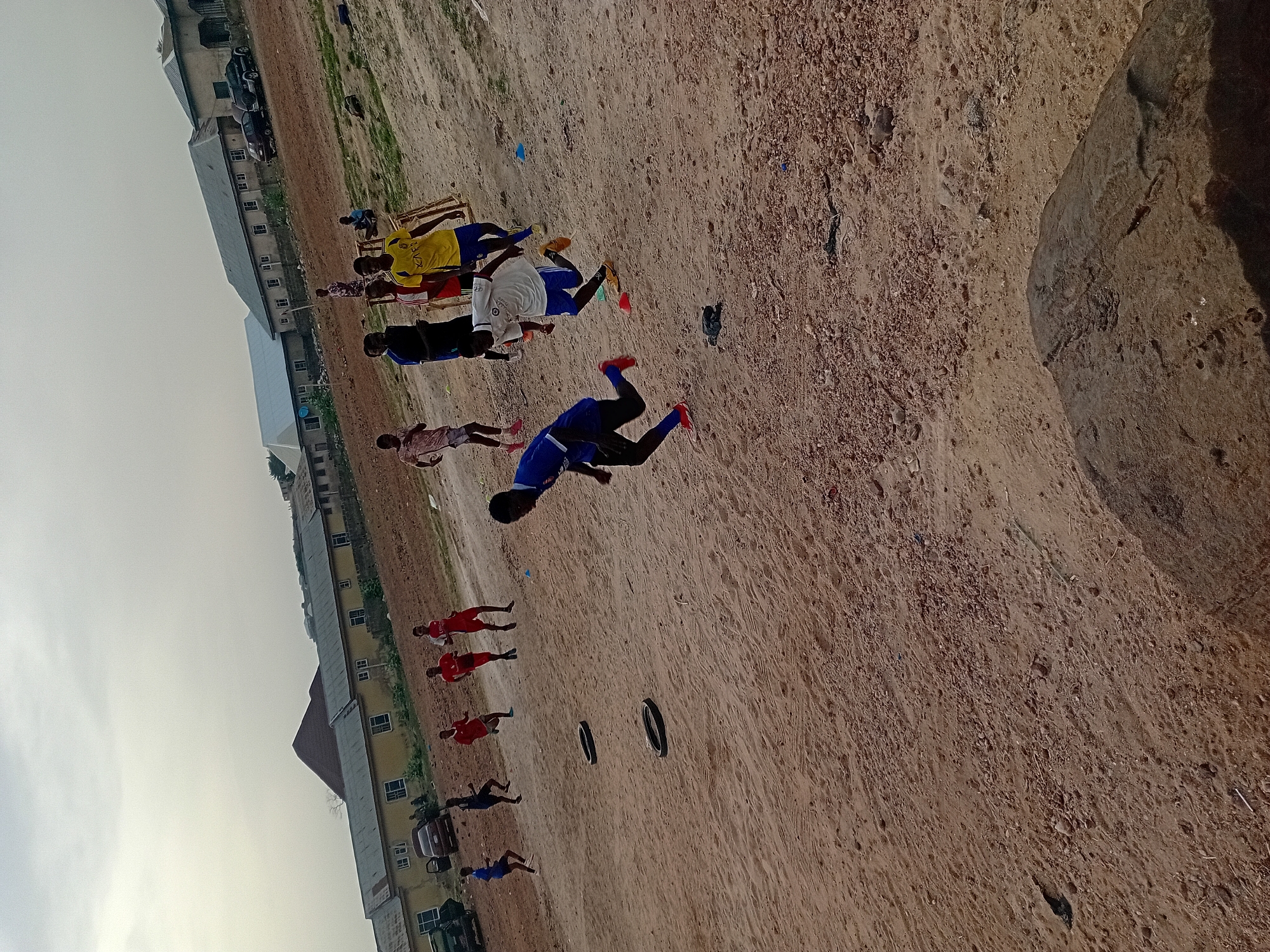 This media file captures an outdoor scene on a dusty, unpaved dirt field, likely serving as a makeshift sports ground. The sky is overcast, indicating a cloudy day with diffused light. In the background, several low-rise buildings line the horizon, suggesting an urban or semi-urban environment.
The primary subjects are numerous individuals, mostly young males, scattered across the field. They are dressed in various athletic uniforms, including blue, red, yellow, and white jerseys, consistent with soccer (football) attire. In the mid-foreground, a prominent figure in a blue jersey, black shorts, and red cleats is captured mid-stride, running towards the right side of the frame. A group of several individuals, including one in a yellow jersey with faint, partially visible text (possibly "XAFIZ" or "KAFU"), stands closer to the background. Other groups and individuals are visible further back and to the left, some also wearing red jerseys.
On the ground, two black tires are laid flat, possibly used as training markers, along with smaller blue and yellow markers. Wooden structures, resembling benches or makeshift goals, are visible in the background near some players. Cars are parked near the buildings in the distance. The overall activity depicted appears to be a soccer practice or an informal game, given the number of players, their sportswear, and the training equipment. The ground itself is reddish-brown dirt, with scattered small stones and debris, and a darker, raised mound is visible on the far right.