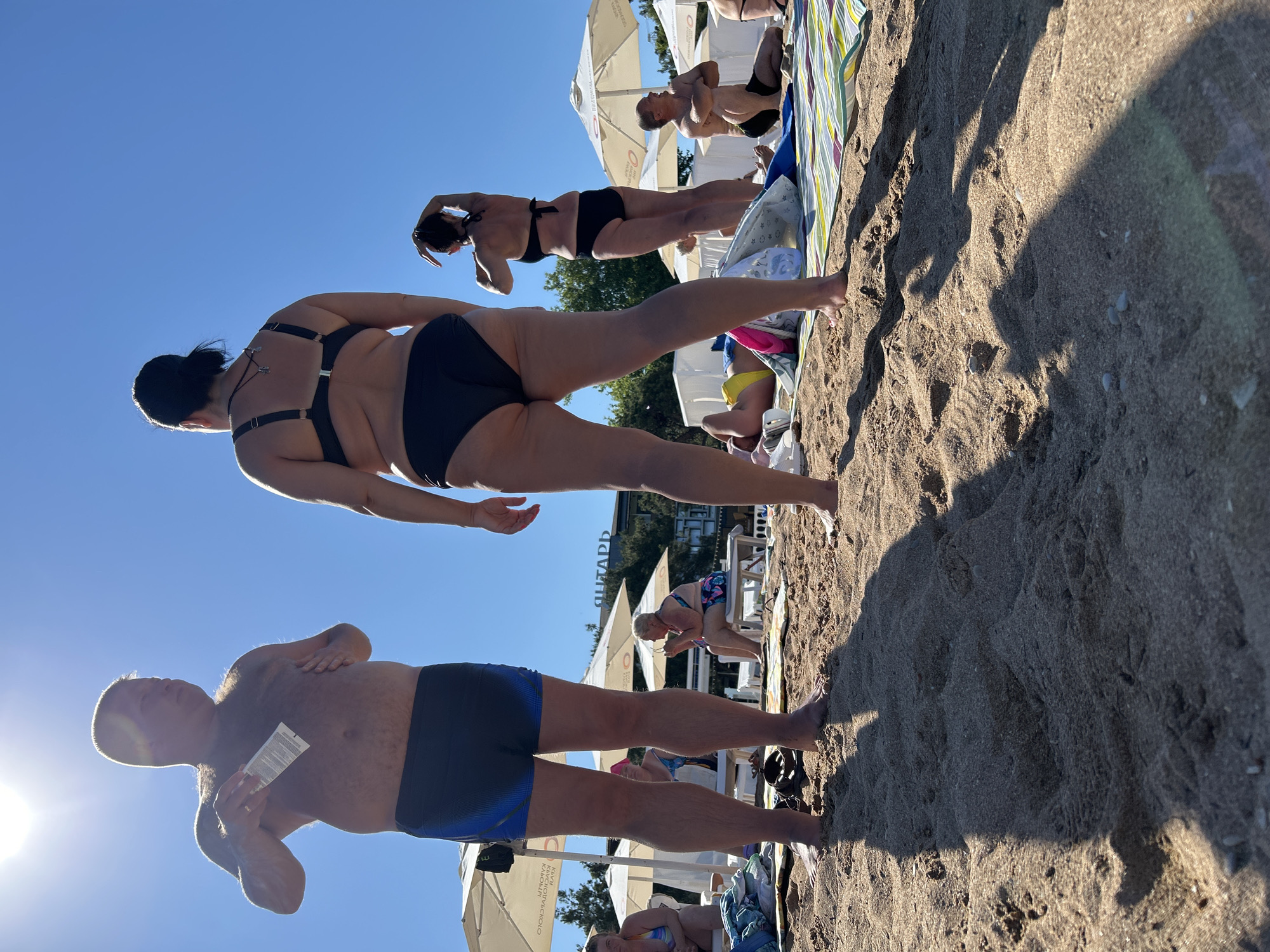 On a sunny day at a beach in Gelendzhik, Russia, a man in black swim trunks stands on the sand, holding a tube of sunscreen and applying it to his chest. Facing away from the camera, a woman in a black bikini stands to his right. In the background, other beachgoers are visible relaxing on loungers under white umbrellas. The sand is light brown, and the sky is a clear blue.