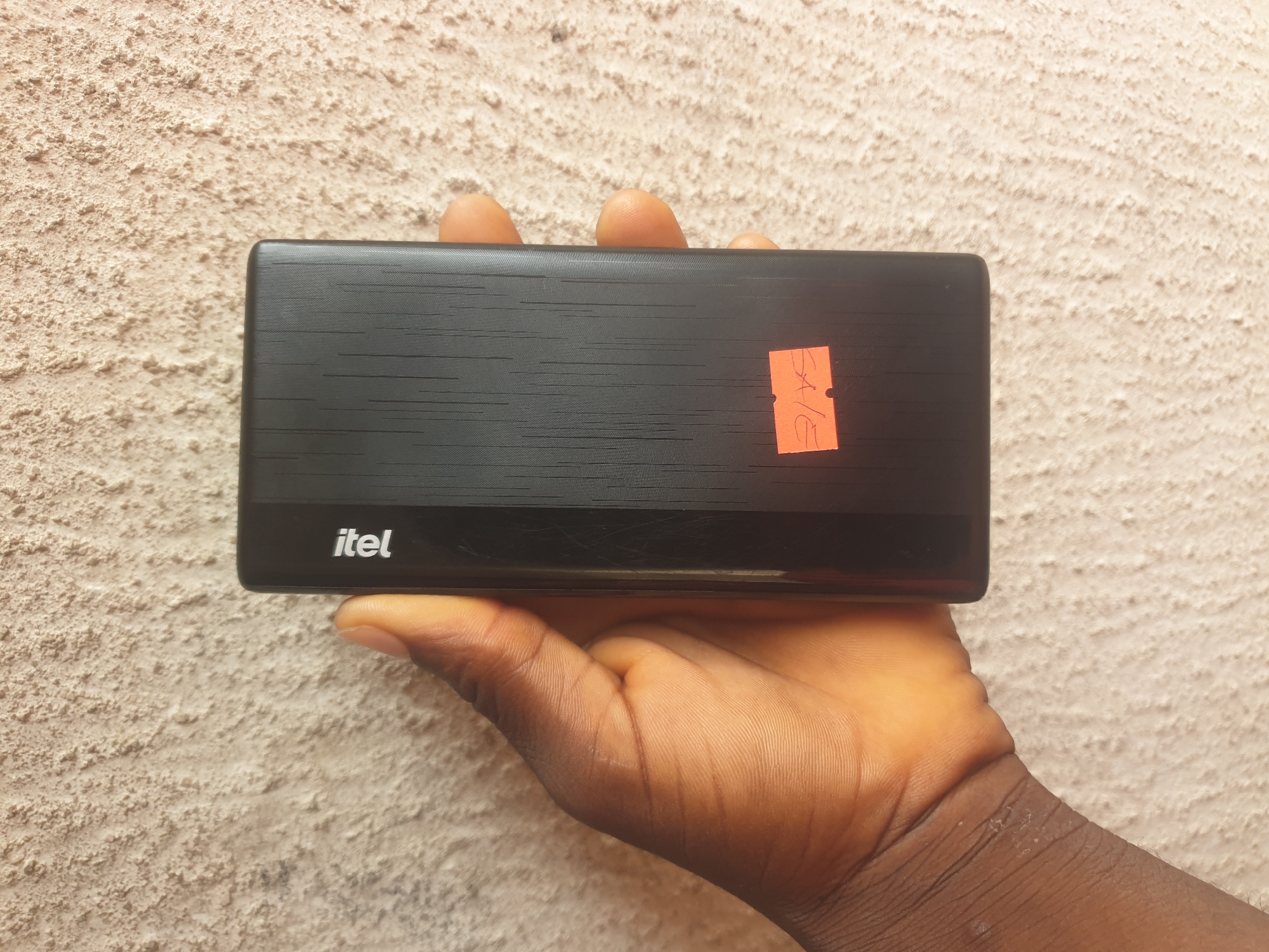 An overhead shot shows a dark-colored power bank held by a person's hand against a textured, pale wall. The power bank has a vertical ribbed texture on its surface and the brand name "itel" is visible in white lettering along the top edge. An orange sticker with handwritten text that appears to be "SALE" is affixed to the lower portion of the power bank. The background is a rough, light-colored wall, typical of many buildings in Jalingo, Nigeria. The lighting suggests an outdoor setting.