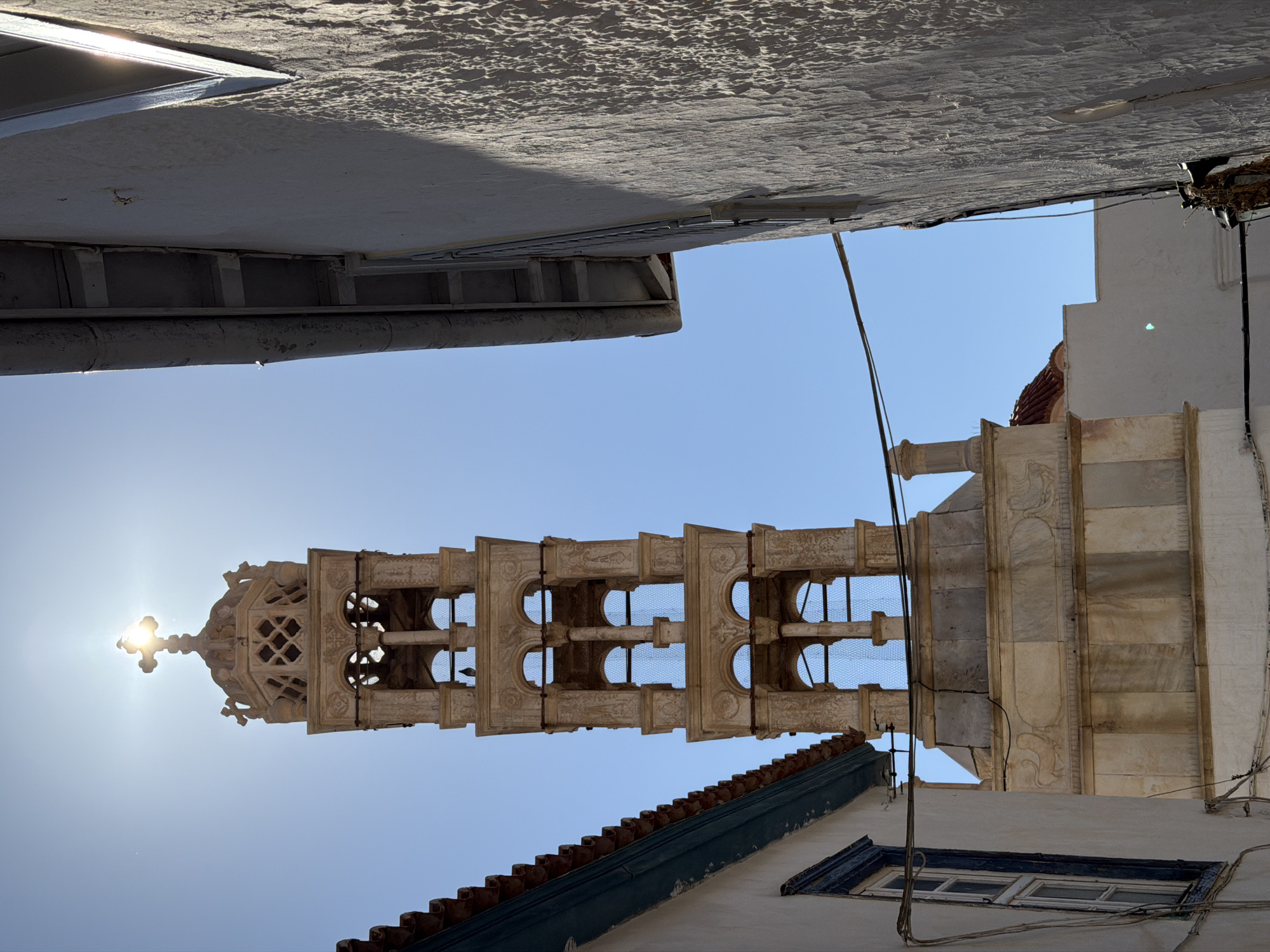 The image shows the bell tower of the Dormition of the Virgin Mary Cathedral in Hydra, Greece, viewed from below at an acute angle. The tower, constructed of light-colored stone, features a series of archways and decorative elements leading to a cross at its apex, where the sun shines through. Buildings frame the top and bottom of the image. Above the tower, there's a section of a whitewashed building or structure. Below, there is a building with a dark red tiled roof and a window with a blue frame. Overhead electrical wires intersect the view of the tower. The sky is a clear blue.