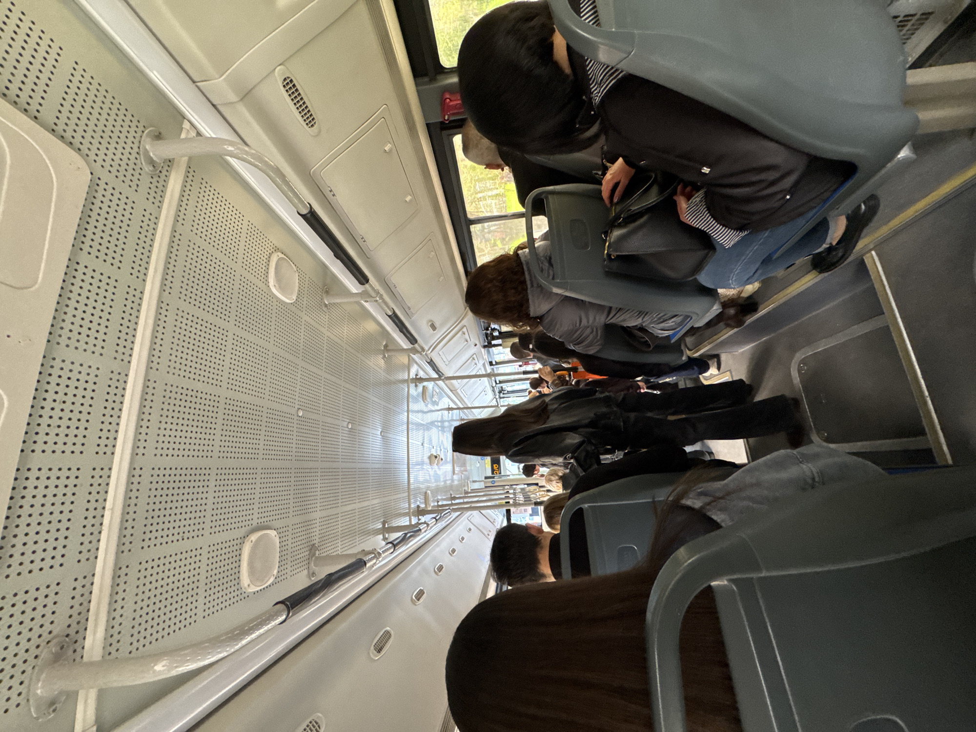 The image depicts the interior of a crowded public transportation vehicle, likely a bus or a tram, with multiple passengers seated. The view is from the rear of the vehicle, looking towards the front. The interior is brightly lit, revealing gray seats, handrails, and a perforated metal wall on the left side. Passengers are of varying ages and genders, with a focus on the backs of their heads and shoulders. Some are wearing jackets and carrying bags. The windows provide a glimpse of the exterior, revealing greenery, suggesting a location within Yerevan, Armenia.