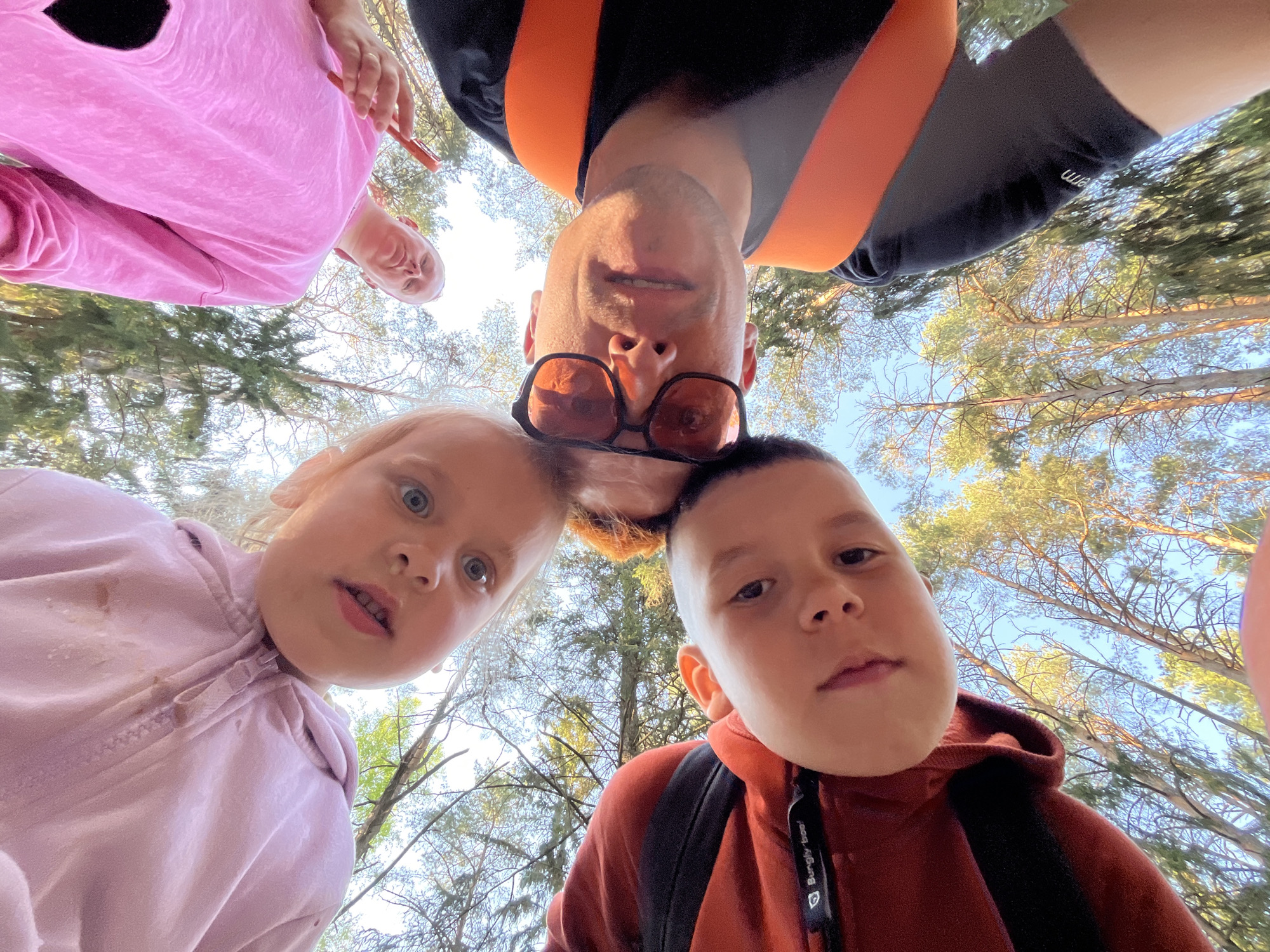 A low-angle shot looking up at four individuals in what appears to be a forest setting, potentially in Chaikovsky, Russia, given the context. At the bottom left, a young girl with fair hair and blue eyes wears a pink hooded jacket with stains. Adjacent to her, a young boy in a reddish-brown hoodie and backpack stares directly at the camera. Above them, an adult male with short ginger hair and aviator sunglasses looks down at the group. He wears a black shirt with orange straps or a harness-like garment. To the upper left, a person in pink loungewear holds a small red object. Trees with sparse leaves fill the background against a clear blue sky. The lighting is bright and even, indicating daylight.