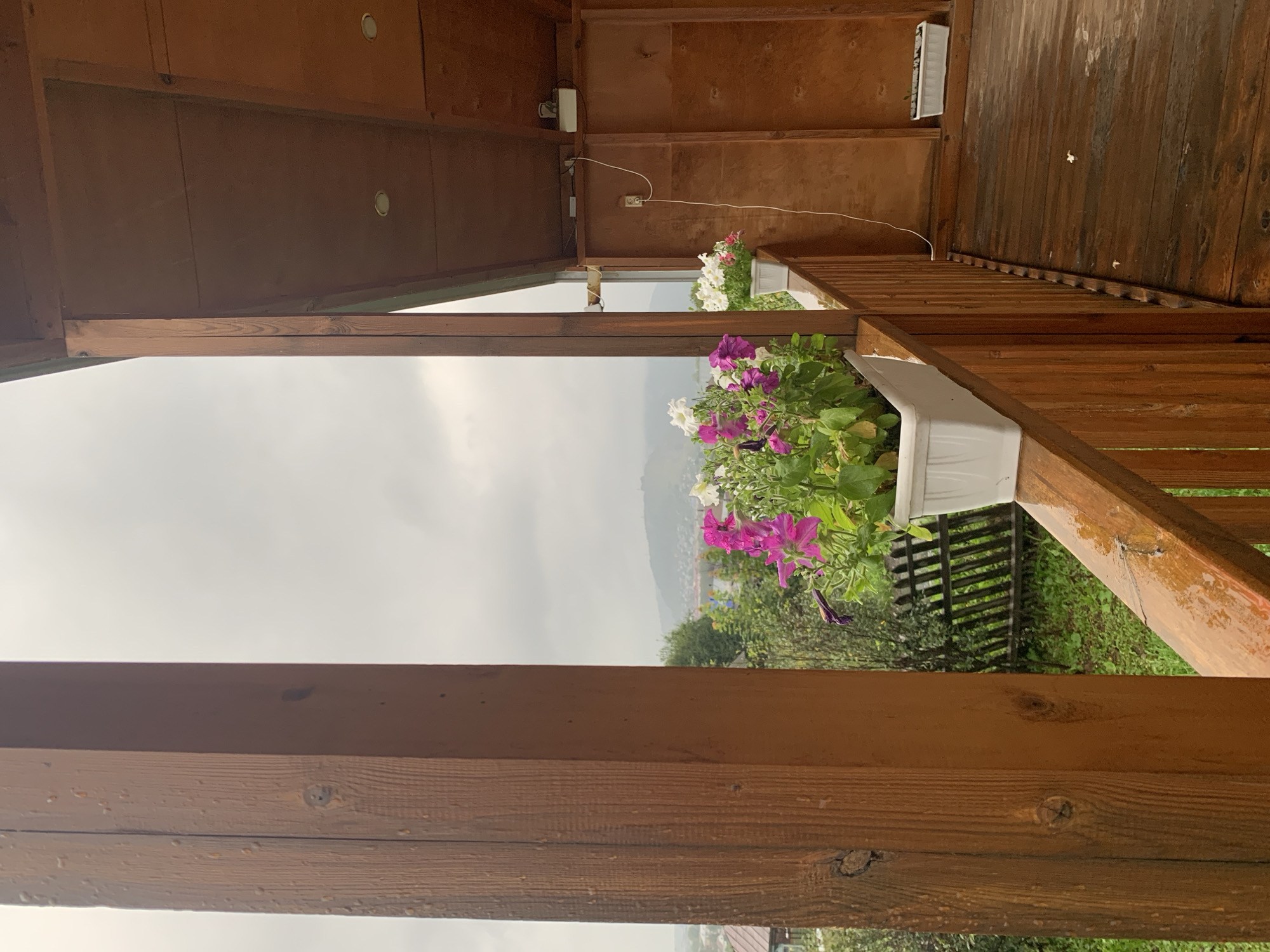 This is a medium shot, taken from a wooden porch overlooking a town. The porch has a wooden railing and floorboards, both appearing wet, suggesting recent rain. On the railing, there are two white planters filled with flowering plants, primarily pink and white petunias. In the background, a town with houses and green vegetation is visible, nestled among rolling hills under a cloudy sky. The architecture suggests a rural or semi-rural setting. The image appears to be taken in Asha, Russia, given the context provided. There are no people or animals present in the image. The scene is static and depicts a view from a covered outdoor space.