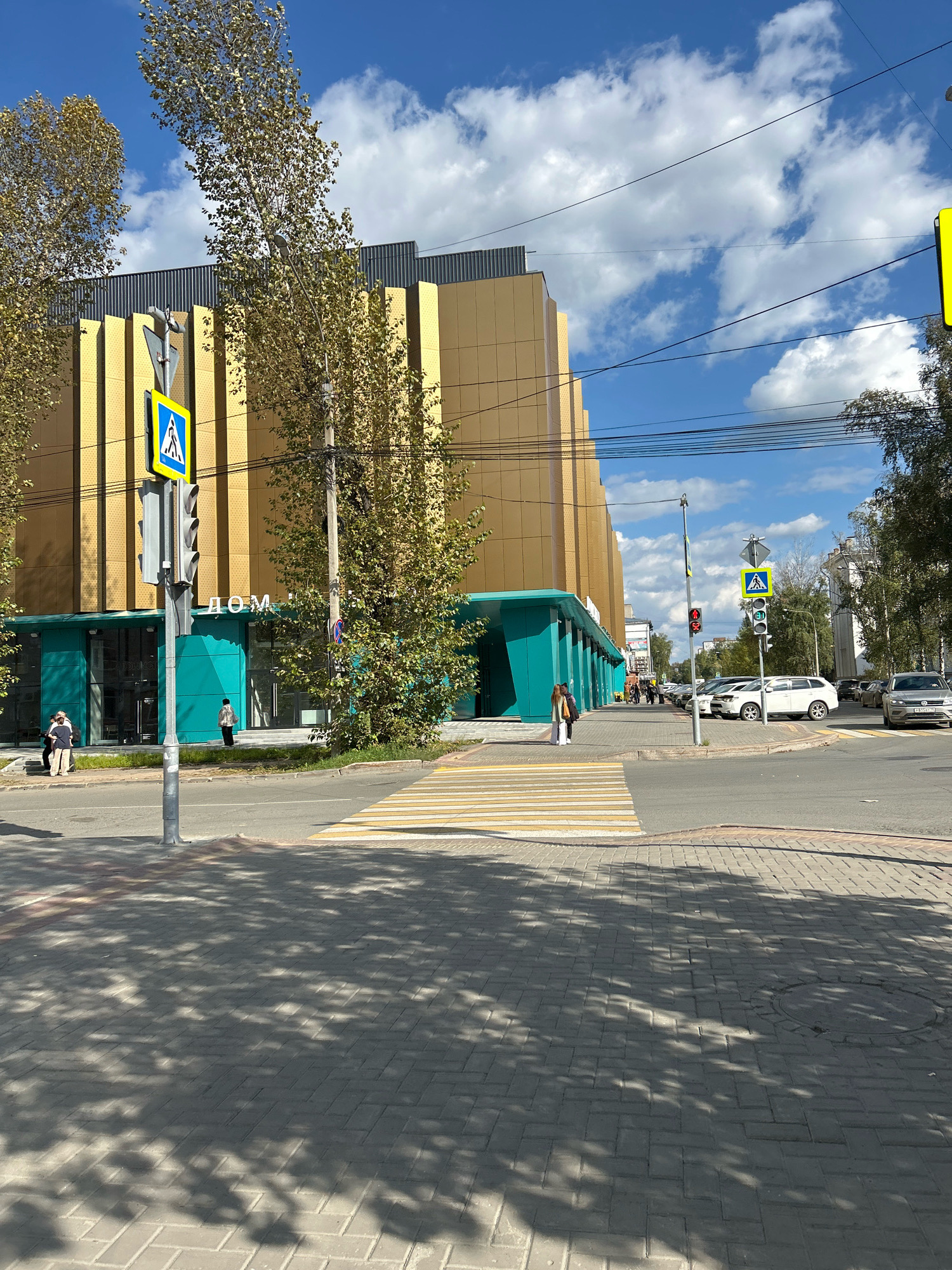 In Tomsk, Russia, a wide, paved street runs in front of a modern, multi-story building with a striking gold facade and teal-colored ground-floor accents. A pedestrian crossing, marked with white and yellow stripes, is visible in the foreground, with tree shadows dappling the pavement. On the left side of the street, a traffic light pole displays a pedestrian crossing sign and a red pedestrian signal. Several individuals are present: two women standing on the sidewalk near the building entrance, and another person walking across the street. A few cars are parked along the right side of the street, and trees line the sidewalk and the building's facade. The sky above is a vibrant blue, dotted with fluffy white clouds.