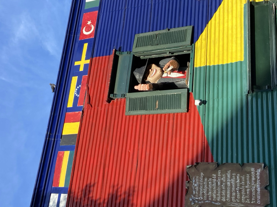 A colorful building exterior features vertical stripes of flags along the left side and bold fields of blue, red, yellow, and green across its facade. A green-framed window reveals a cartoonish figure in a dark suit, seemingly smoking a cigar. To the right, another window is partially open. Below the window with the figure, a weathered plaque displays Spanish text describing "Venerado Maestro Quinquela" and evokes imagery of the Riachuelo river and a sun-soaked neighborhood.