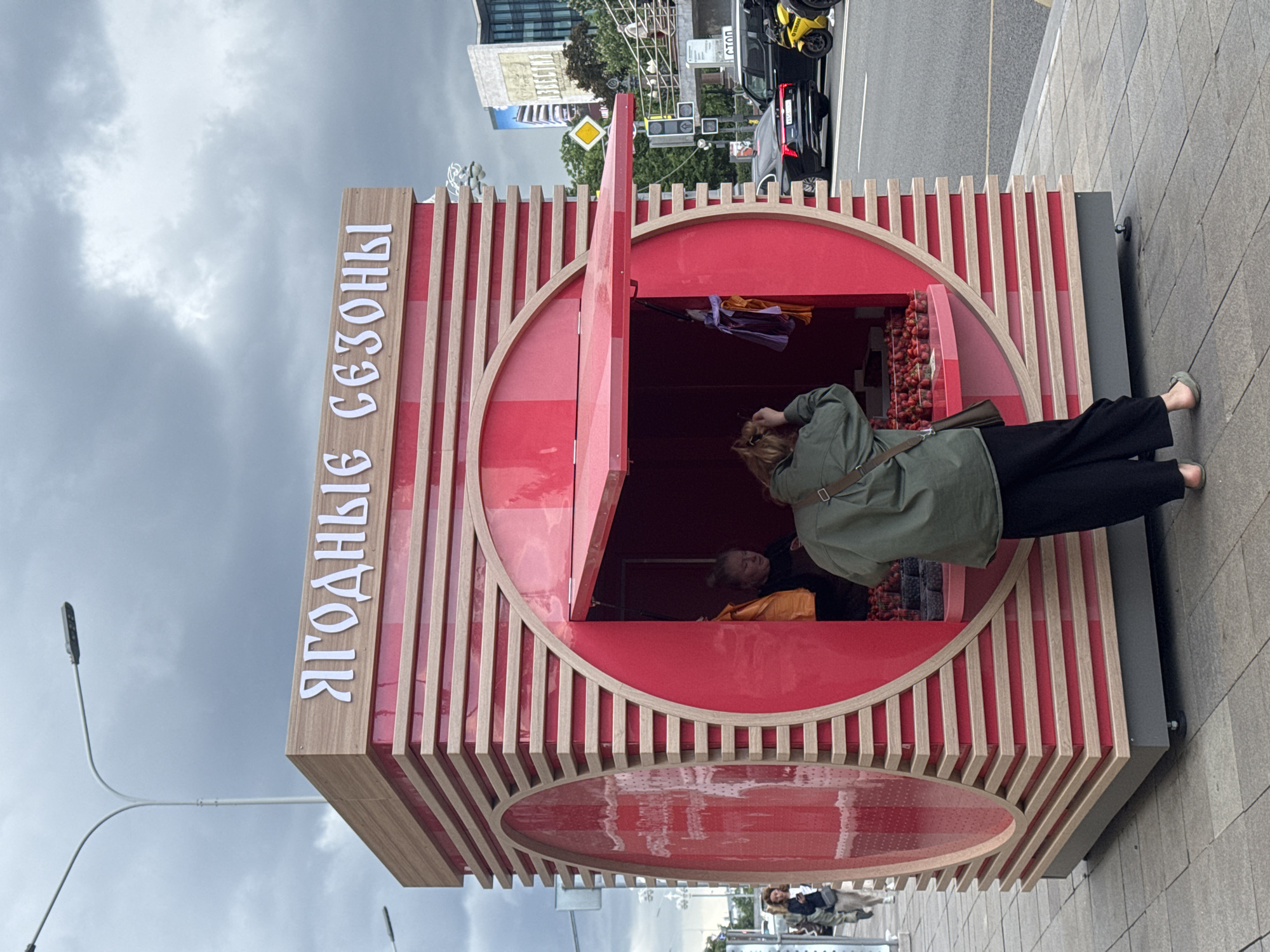 The image shows a food kiosk on a city street. The kiosk is a red and wood structure with a circular window and an open red door. Text in Cyrillic script is visible on the side of the kiosk. A woman in a green jacket and black pants is bending down to look inside the kiosk. Inside the kiosk, a person in an orange top is visible. Behind the kiosk is a city street with cars and buildings. The sky is overcast. Based on the Cyrillic text "ЯГОДНЫЕ СЕЗОНЫ" (Berry Seasons), which is a known brand, and the architectural style in the background, this image is located in Moscow, Russia.