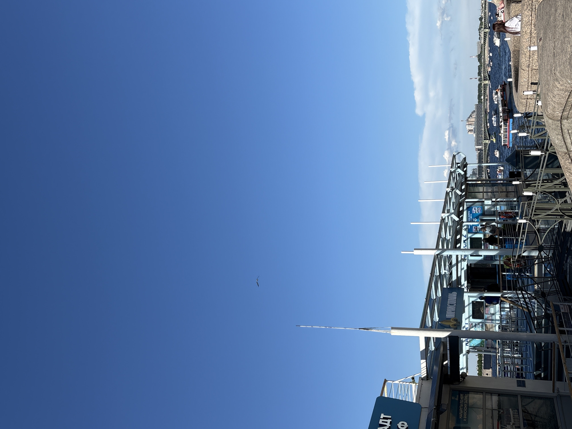 The image captures a detailed scene from a pier on the Neva River in Saint Petersburg, Russia, under a clear blue sky.
In the immediate foreground, a modern pier structure is visible, featuring a light-colored framework, transparent roofing, and multiple white vertical poles. On the left, a blue sign displays Russian text, partially obscured, with visible characters "АДТ" and below it, "МАРШРУТЫ ПО НЕВЕ И ФИНСКОМУ ЗАЛИВУ" (Routes along the Neva and the Gulf of Finland), confirming the location and purpose. Another prominent sign on the pier reads "ПРИСТАНЬ" (Pier). Several individuals are present on the pier and its associated walkways; some are near what appear to be ticket booths or service windows. Metal railings and barriers define the pedestrian areas. To the right, a sturdy stone embankment with a curved top provides a viewing point. A person with dark hair, wearing a white top, is seated on this embankment, facing away from the camera and towards the river.
The midground is dominated by the wide expanse of the Neva River, colored a deep blue. Numerous boats of various types, including motorboats and small passenger vessels, are scattered across the water. Many boats are in motion, creating visible wakes, while others appear to be stationary. In the distance, a large bridge with a green structural framework spans the river. Beyond the bridge, the cityscape of Saint Petersburg is visible, featuring various buildings, including a distinctive multi-tiered structure with a unique roofline on the far bank.
The background consists primarily of a bright, clear blue sky, occupying the upper two-thirds of the frame. A single, small, dark bird is captured in flight in the upper left quadrant of the sky. A few sparse, white cumulus clouds are visible along the horizon line, particularly towards the right side of the image. The overall lighting suggests a sunny day with excellent visibility.