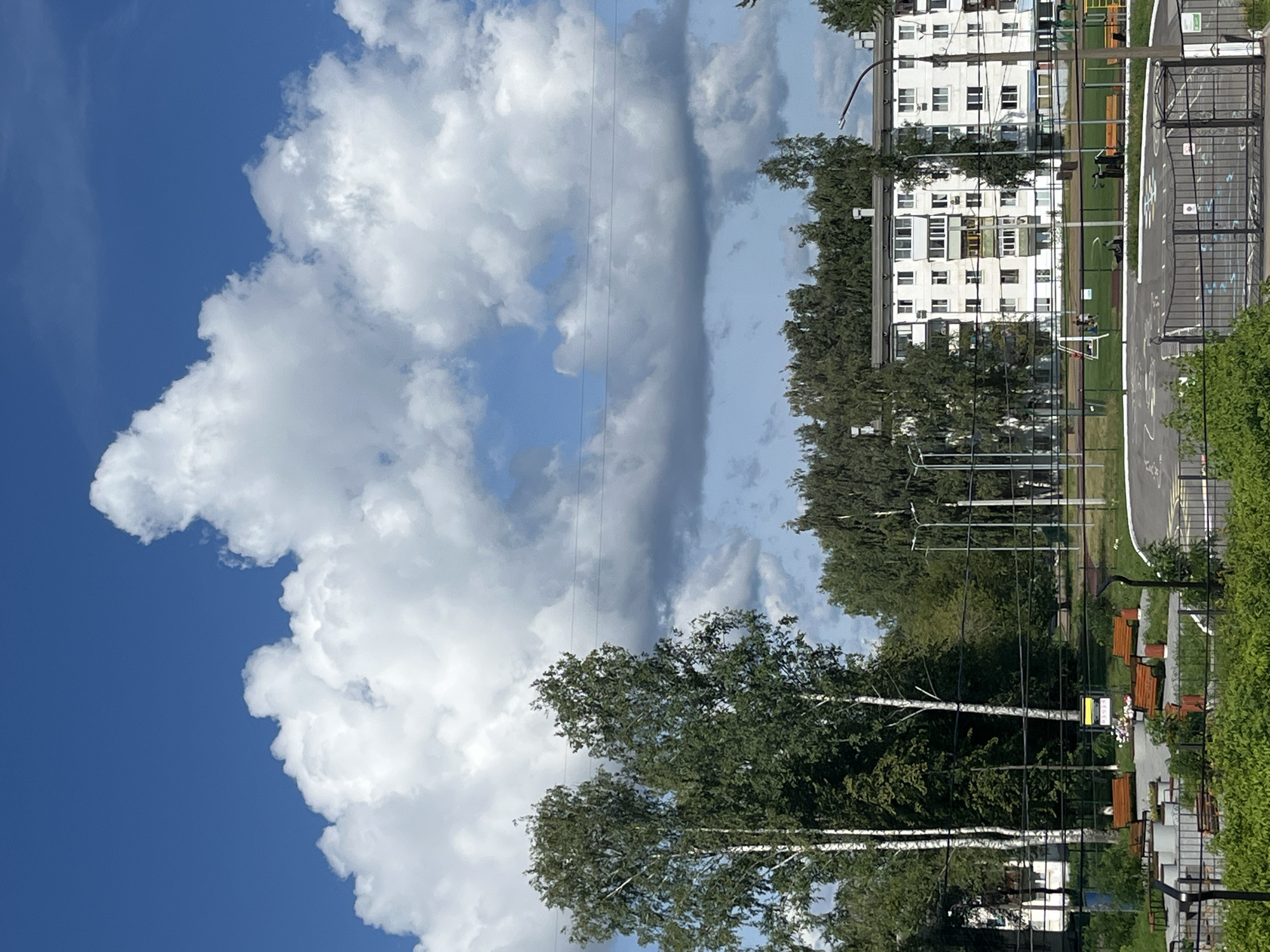 The image captures a bright, sunny day in Chaikovsky, Russia, with a dramatic sky dominated by large, fluffy white cumulus clouds against a deep blue background. In the foreground, several trees with thin white trunks and dense green foliage are prominent, suggesting a park or residential area. Beyond the trees, a multi-story white apartment building with visible windows and balconies stands, indicative of Soviet-era architecture. The building is surrounded by a line of trees. In front of the building, a playground area with some equipment and benches is visible. A paved circular area with markings for games is also present, bordered by a metal fence. Several people are scattered throughout the scene, including two figures near a swing set on the playground and others in the distance. The overall impression is of a typical urban summer day in a residential district, with elements of nature and community spaces.