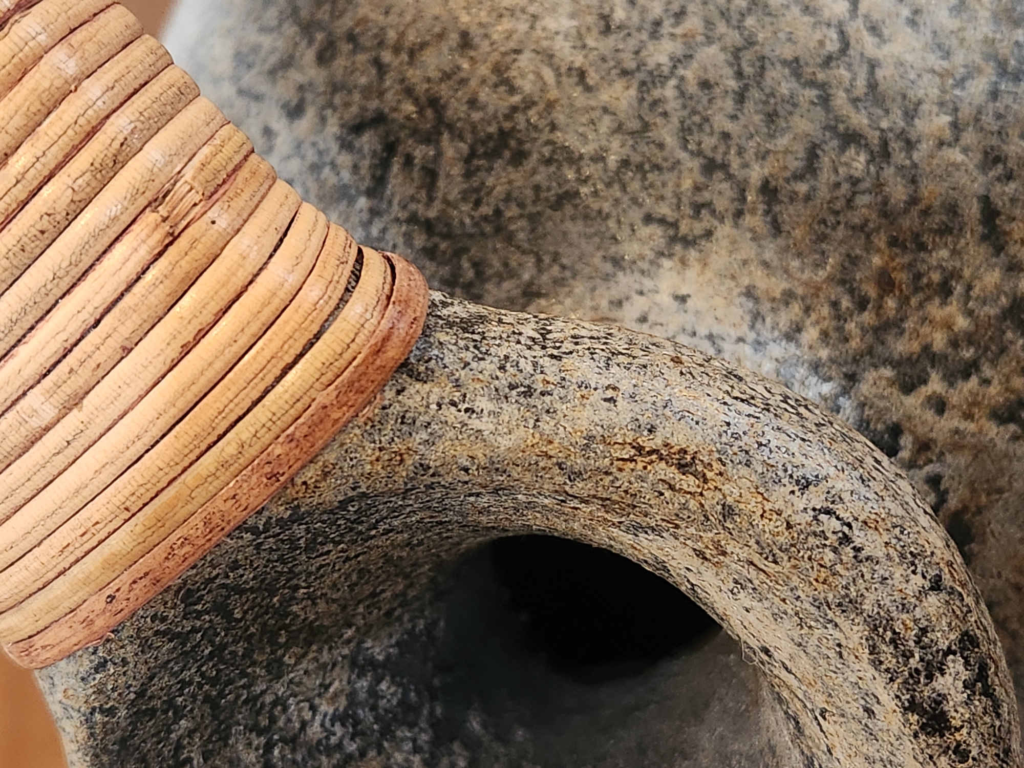 The image is a close-up, slightly tilted shot of a textured, weathered object, likely a stone vessel with a woven handle. The stone has a rough, granular surface with variations in color, ranging from light grey to dark speckles. A distinct, circular opening is visible in the lower left portion of the frame. The handle, made of tightly woven, light-colored material, possibly bamboo or wicker, is positioned on the upper part of the object, resting on the stone. The woven material shows individual strands with a natural, aged appearance, and some sections appear slightly frayed or cracked. The background is softly blurred, revealing more of the stone's textured surface. There are no people, actions, or interactions depicted. This image does not provide any information about its location in Maitland, United States of America.