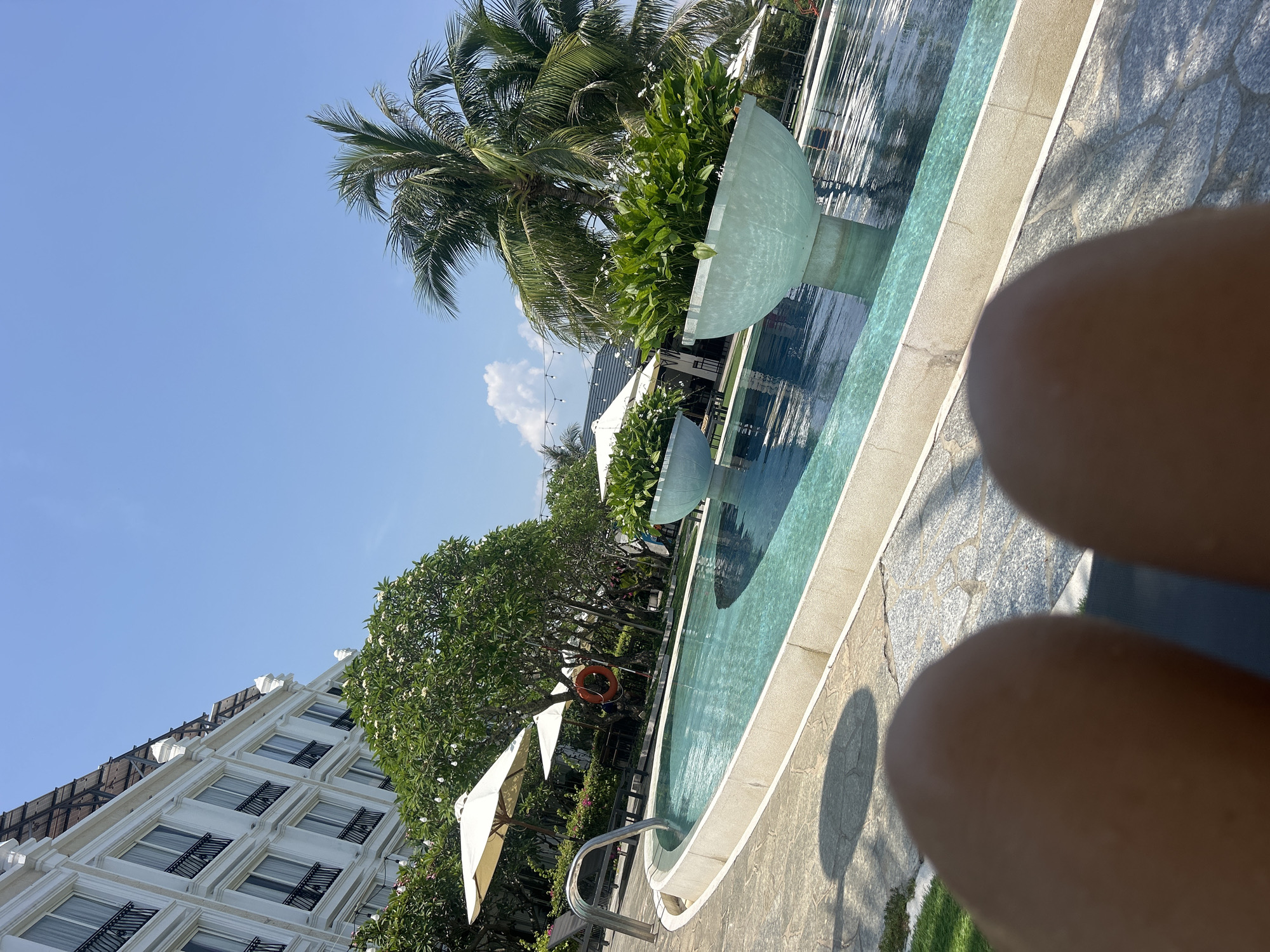 The image shows a swimming pool area at what appears to be a resort or hotel in Bac Nha Trang Ward, Vietnam. The pool is bordered by stone and features decorative bowl-shaped fountains with greenery. White buildings with dark-shuttered windows are visible in the background. Palm trees and sun umbrellas provide shade. The foreground is dominated by what seems to be a person's knees, indicating the photo was taken from a lounging position.