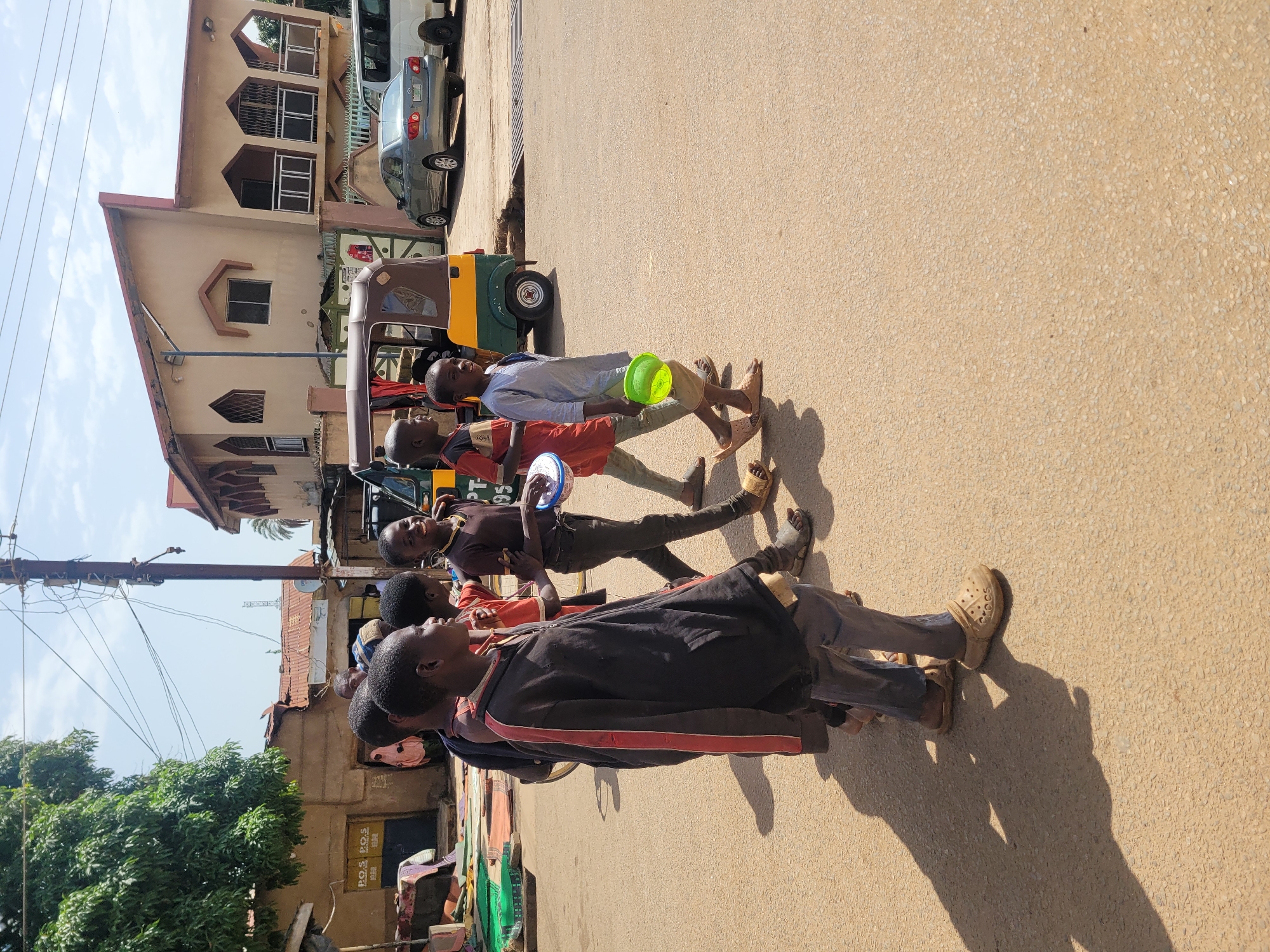 A group of young boys are walking on a dusty asphalt road in Jos, Nigeria. The boys appear to be of school age and are dressed in casual clothing. Some of them are carrying items, including a green plastic bowl. In the background, there are several buildings, some of which appear to be shops. A yellow and green motorized rickshaw, commonly known as a "Keke Napep" in Nigeria, is parked on the side of the road, along with a gray sedan and a white van. Overhead, power lines crisscross the sky. The sun is shining, casting long shadows on the road.