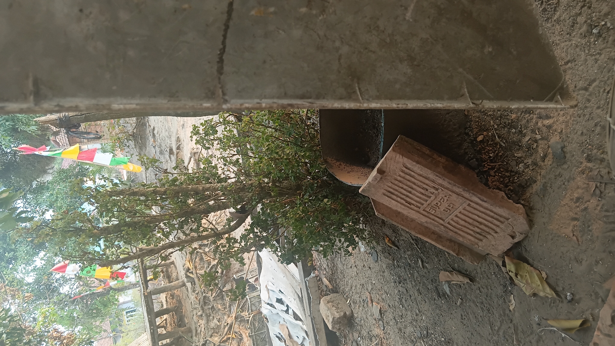 The image depicts an outdoor scene in what appears to be a rural or semi-rural area, possibly on the outskirts of Prabumulih, Indonesia. In the foreground, a broken brick with raised lettering, possibly a brand name, lies on the dusty ground. To the right, a large, rough concrete pillar partially obscures the view. Behind the pillar and to the left, a green, leafy bush is prominent, with several trees visible in the background. Colorful flags, red, white, yellow, and green, are strung between poles and branches, suggesting a festive or commemorative occasion. In the mid-ground, remnants of what might be a collapsed structure or debris are visible, including corrugated metal sheets and wooden supports. The ground is uneven and appears to be a mix of dirt, sand, and scattered debris. The overall impression is of an undeveloped or partially developed area, possibly undergoing construction or experiencing disrepair. No people are visible in the image.