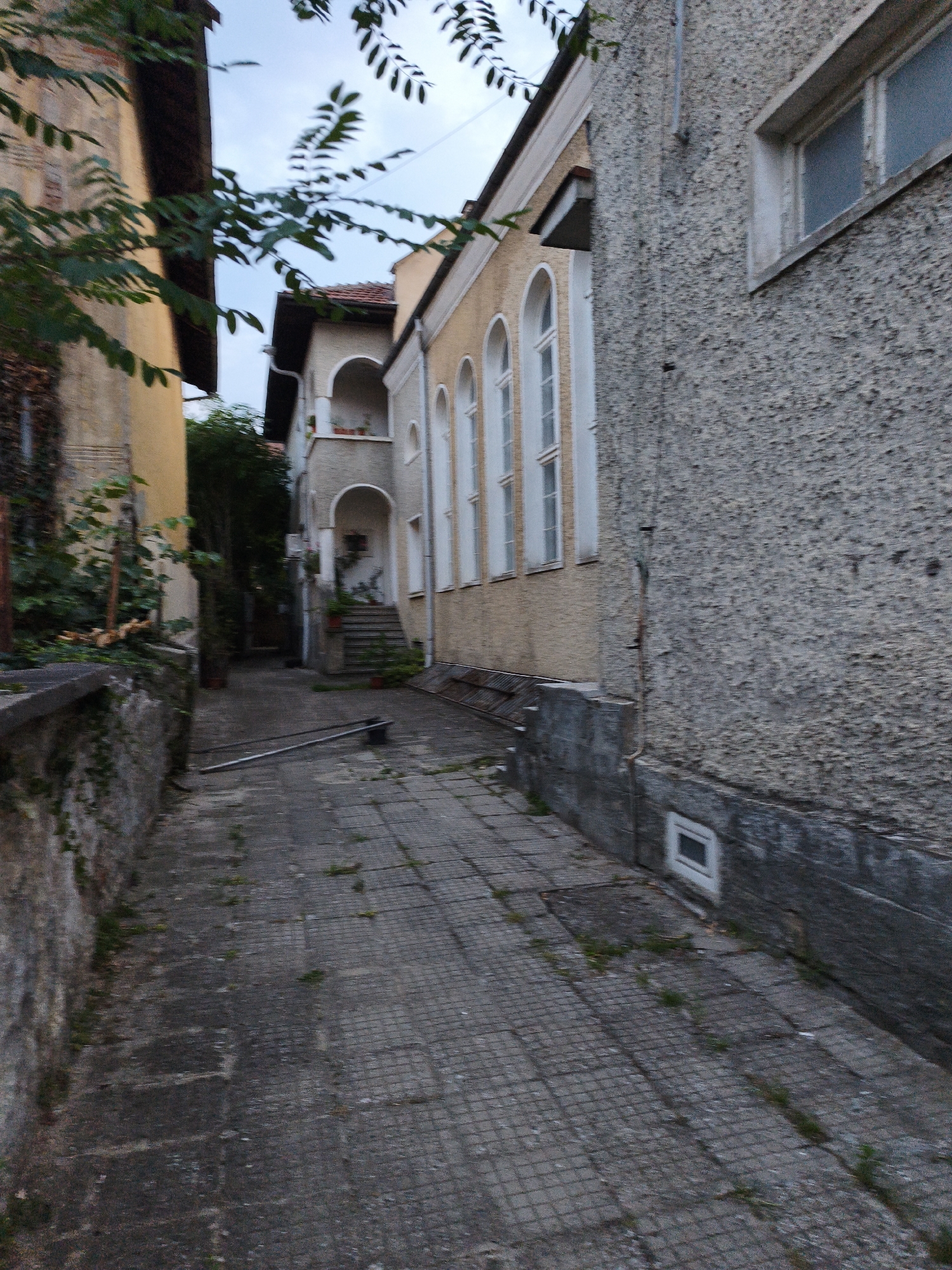 An exterior view of a narrow alleyway in Lovech, Bulgaria, with buildings on either side. The alley is paved with gray tiles. On the left, a textured stone wall is partially covered with ivy. The building on the right has a light-colored stucco exterior with several arched windows with white frames. A second building, visible in the background, has a pale yellow facade with similar arched windows and a red-tiled roof. The scene is dimly lit, suggesting it may be early morning or late evening. There are no people or animals visible in the image.