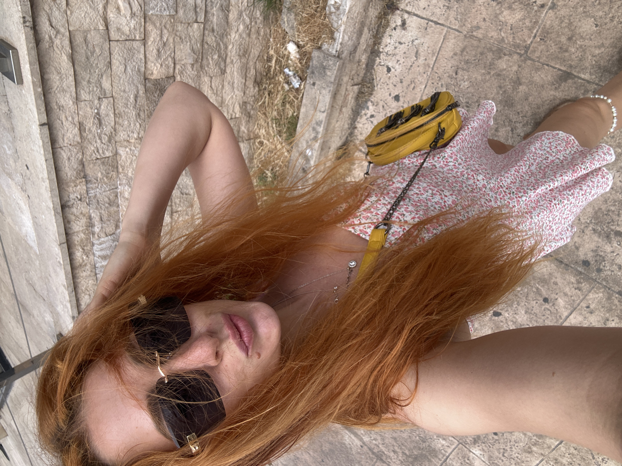 This high-angle shot captures a person lying down or leaning back outdoors, with the camera positioned directly above their face. The subject has long, flowing reddish-blonde hair that frames their face and spreads over their chest and shoulders. They are wearing dark sunglasses with gold accents, obscuring their eyes, and their lips are slightly parted, revealing a natural pink color. A small mole is visible below the left corner of their mouth. A delicate necklace with small, silvery charms adorns their neck.
The person is wearing a light-colored dress with a small pink and white floral pattern. A bright yellow cross-body bag, detailed with fringes and featuring a chain strap, rests on the dress near their right side. A pale arm is visible in the foreground, and a pearl bracelet is worn on one of the wrists.
The setting is an outdoor area, likely a sidewalk or patio, characterized by textured stone paving in varying shades of light brown and beige. To the left, a vertical stone wall composed of rectangular blocks features a modern light switch or panel. Further in the background, a patch of dry grass, dirt, and some scattered litter, including a small plastic bottle, can be seen. The bright, clear lighting suggests it is daytime. The scene depicts a relaxed moment in an outdoor setting, consistent with areas found in Budva, Montenegro. There is no visible text in the image.