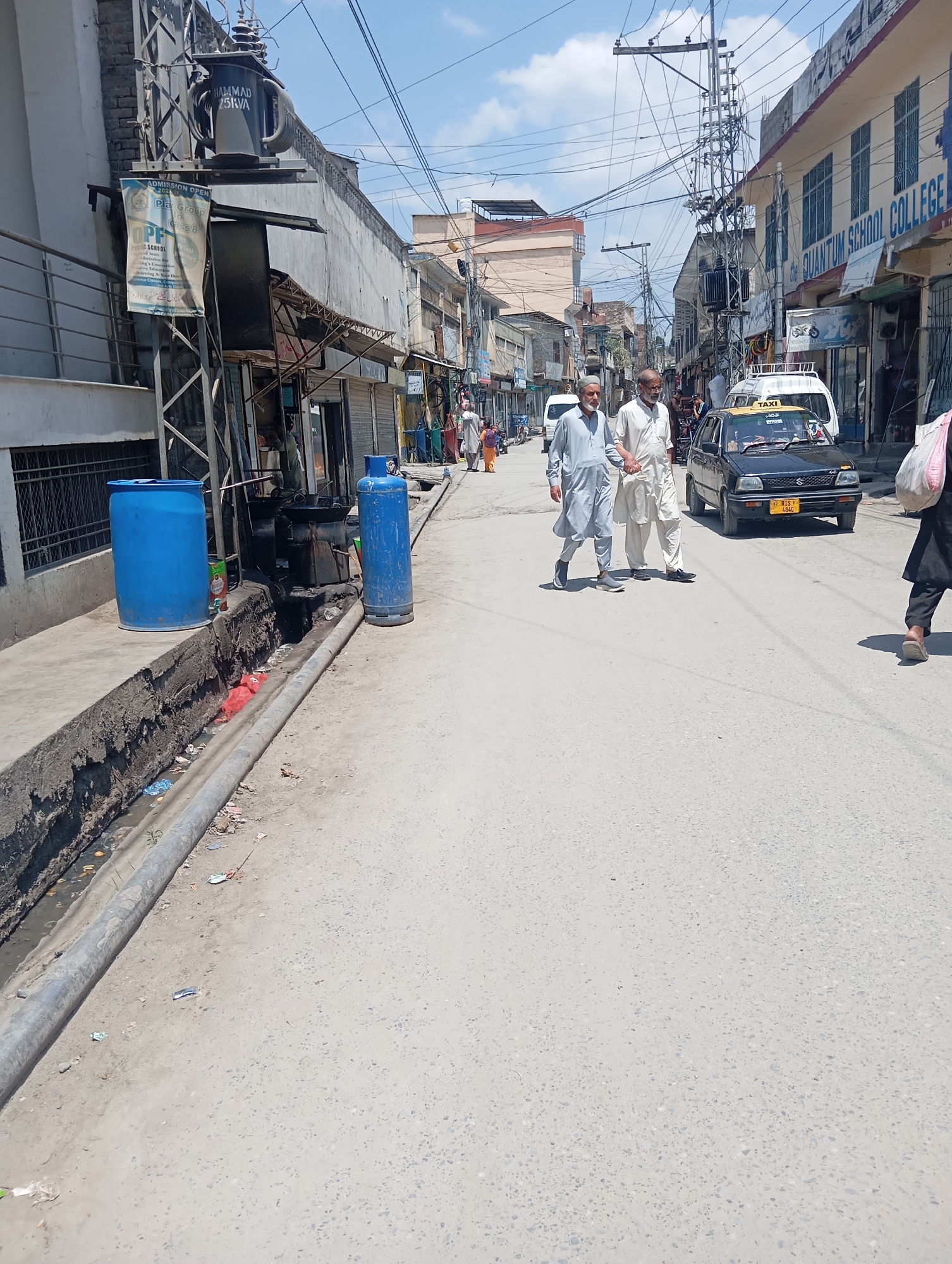 This image depicts a street scene in Mansehra, Pakistan. In the foreground, a grimy, open drainage ditch runs alongside the road. A large blue plastic barrel and a blue propane tank are visible near the ditch. Two men in traditional Pakistani attire are walking across the street, one holding the other's hand. A black car with a "TAXI" sign on its roof is stopped behind them, and a white van is further down the road. The buildings lining the street appear to be a mix of commercial establishments and residences. A prominent sign on one building reads "QUANTUM SCHOOL COLLEGE." Overhead, a complex network of electrical wires crisscrosses the sky, typical of many urban areas in Pakistan. The sky is bright and partly cloudy. The overall impression is one of a busy, lived-in urban environment.