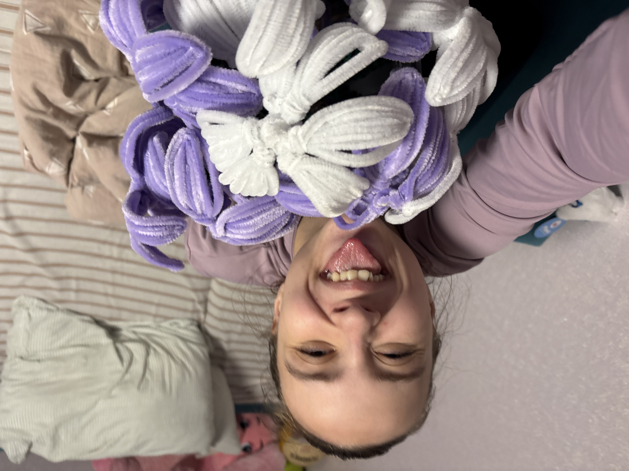A young woman is captured in a playful, upside-down selfie. She is smiling broadly with her tongue sticking out, looking directly at the camera. Her eyes are partially closed in a joyful expression. She wears a light purple long-sleeved top.
She is holding a large cluster of fluffy, ribbon-like bows or hair accessories above her head, which are prominently featured in the foreground. These accessories are primarily in two colors: bright white and a soft lavender purple. They appear to be made from a textured, possibly chenille or pipe cleaner-like material, creating a soft, voluminous effect.
The background suggests an indoor setting, likely a bedroom in Прикамский, Russia. A bed is visible with striped bedding in light brown and beige tones, along with a light-colored, possibly corduroy, pillow. A beige comforter is also present. Some indistinct items, including a pink object (possibly a toy or blanket), are partially visible in the lower part of the frame. The lighting is soft and even, suggesting an indoor environment without strong directional light. There is no visible text in the image. The overall mood is cheerful and lighthearted.