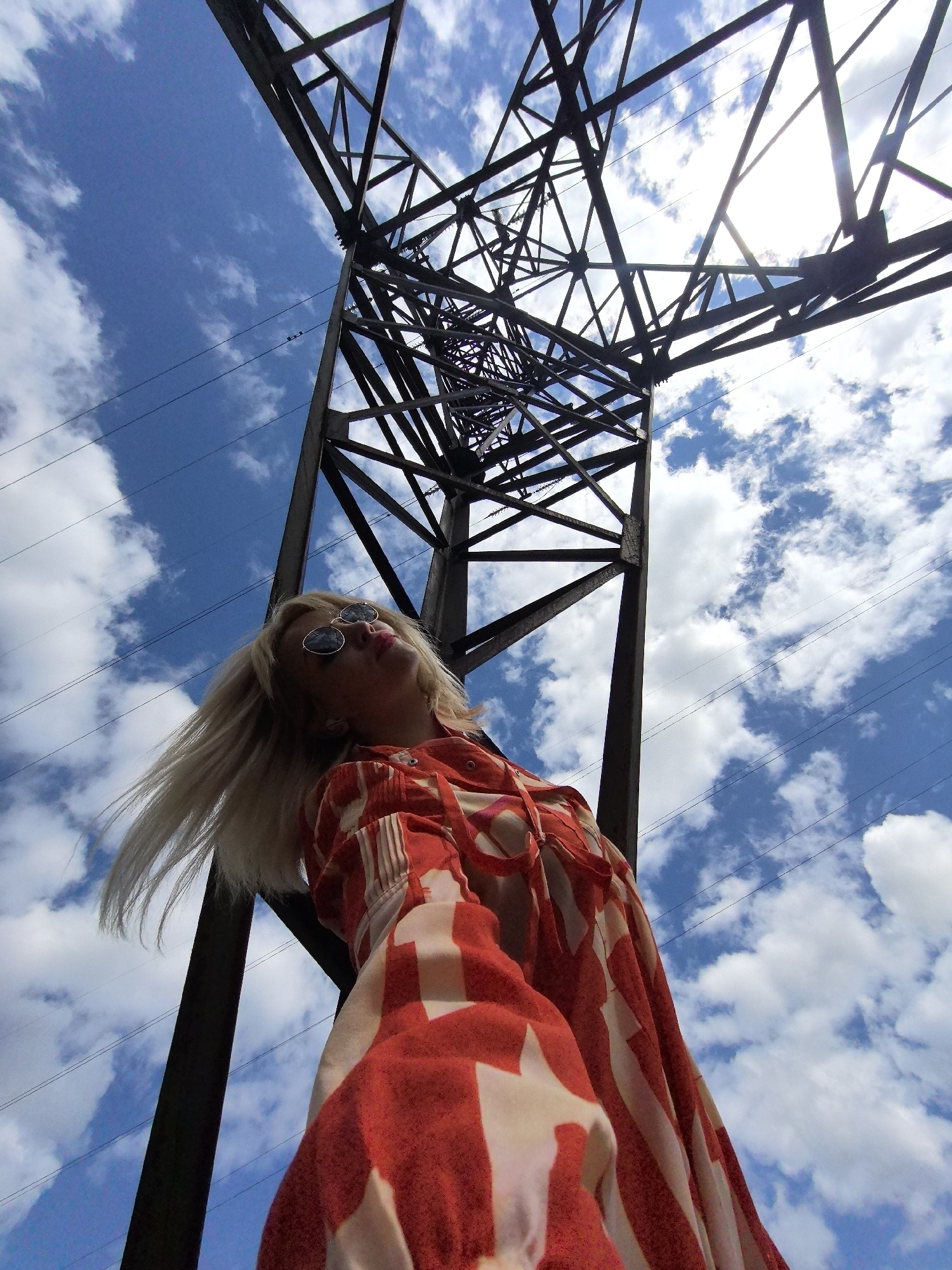An outdoor, low-angle, medium shot captures a blonde woman from the chest up, looking upwards as she stands beneath a towering industrial metal structure against a bright blue sky filled with fluffy white clouds. The woman, positioned on the left side of the frame, wears round, dark-lensed sunglasses and a red and white striped or patterned garment. Her blonde hair is windswept, extending to the left and partially obscuring her face. The large metal structure, likely a power pylon, dominates the upper portion of the image, with its dark, angular beams crisscrossing and extending upwards. Multiple power lines stretch horizontally across the sky. The bright sun is visible through the metal lattice work.