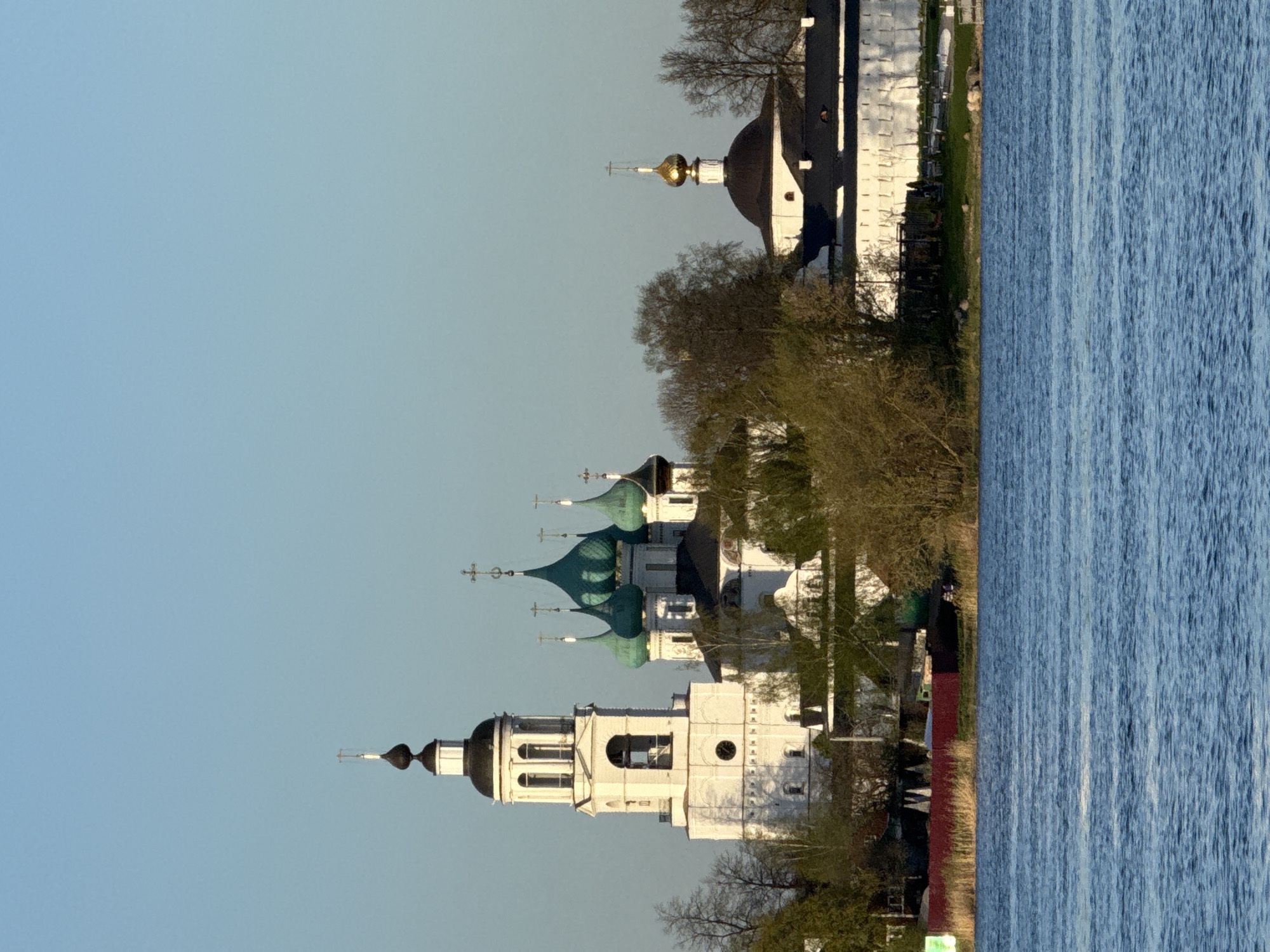 The image depicts a serene landscape with a river on the right side. The sky is a gradient of light blue. On the left side, there is a cluster of white buildings with various domes and crosses, suggesting religious architecture, likely a monastery. These buildings are situated among trees with budding leaves. The scene is indicative of the city of Rusanovo, Russia.