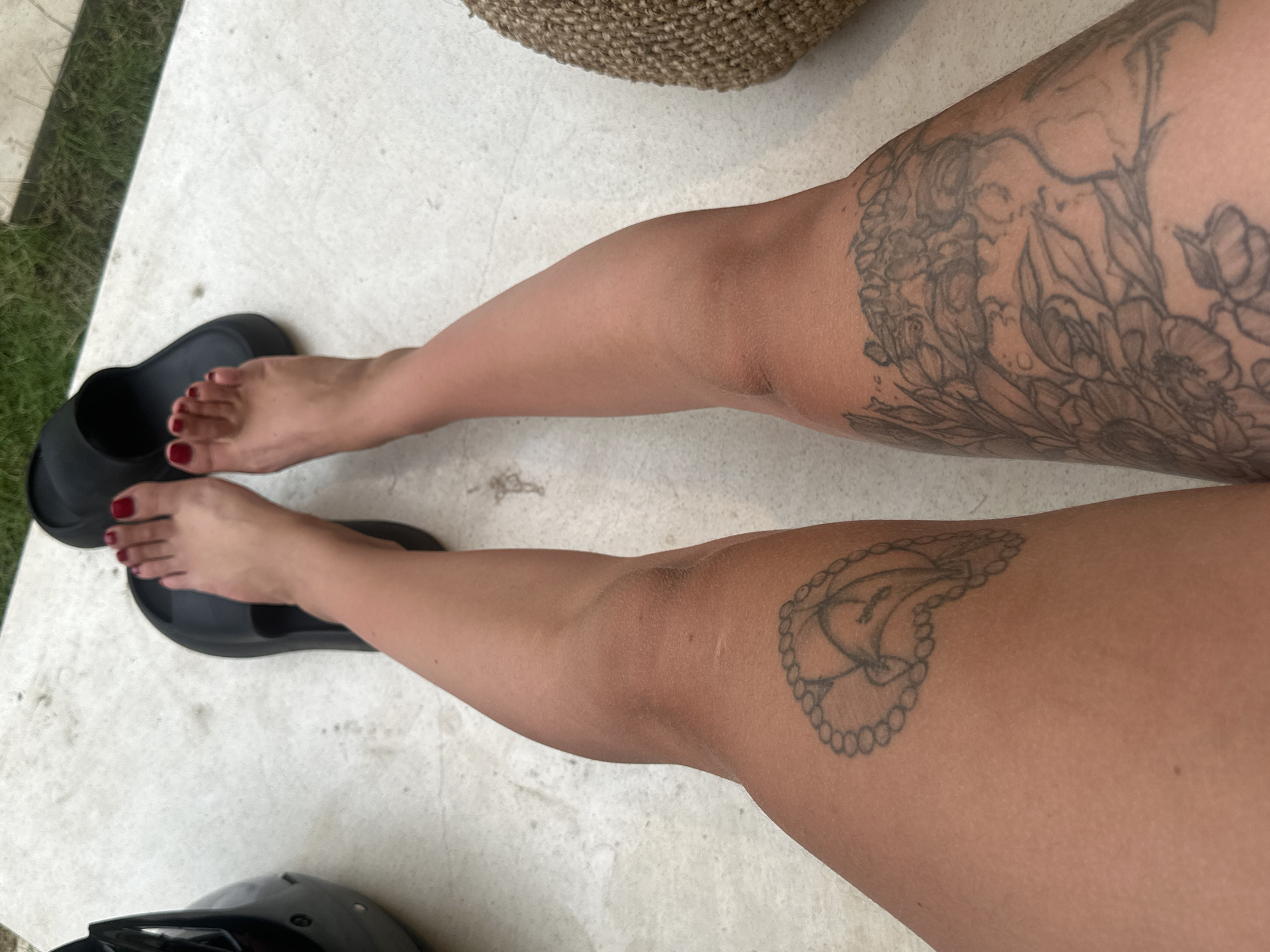The image shows a person's legs and feet, viewed from a slightly elevated angle. The person is wearing black slide sandals and has red nail polish on their toes. There are two tattoos visible on the legs: a small, outline tattoo of a locket with a bird on the left thigh, and a larger, more intricate tattoo of flowers and possibly a face on the right thigh. The person is standing or sitting on a light-colored, speckled surface, possibly concrete or stone, next to some green grass. A black helmet is partially visible to the left of the frame, and a woven basket is to the right. The scene appears to be outdoors.