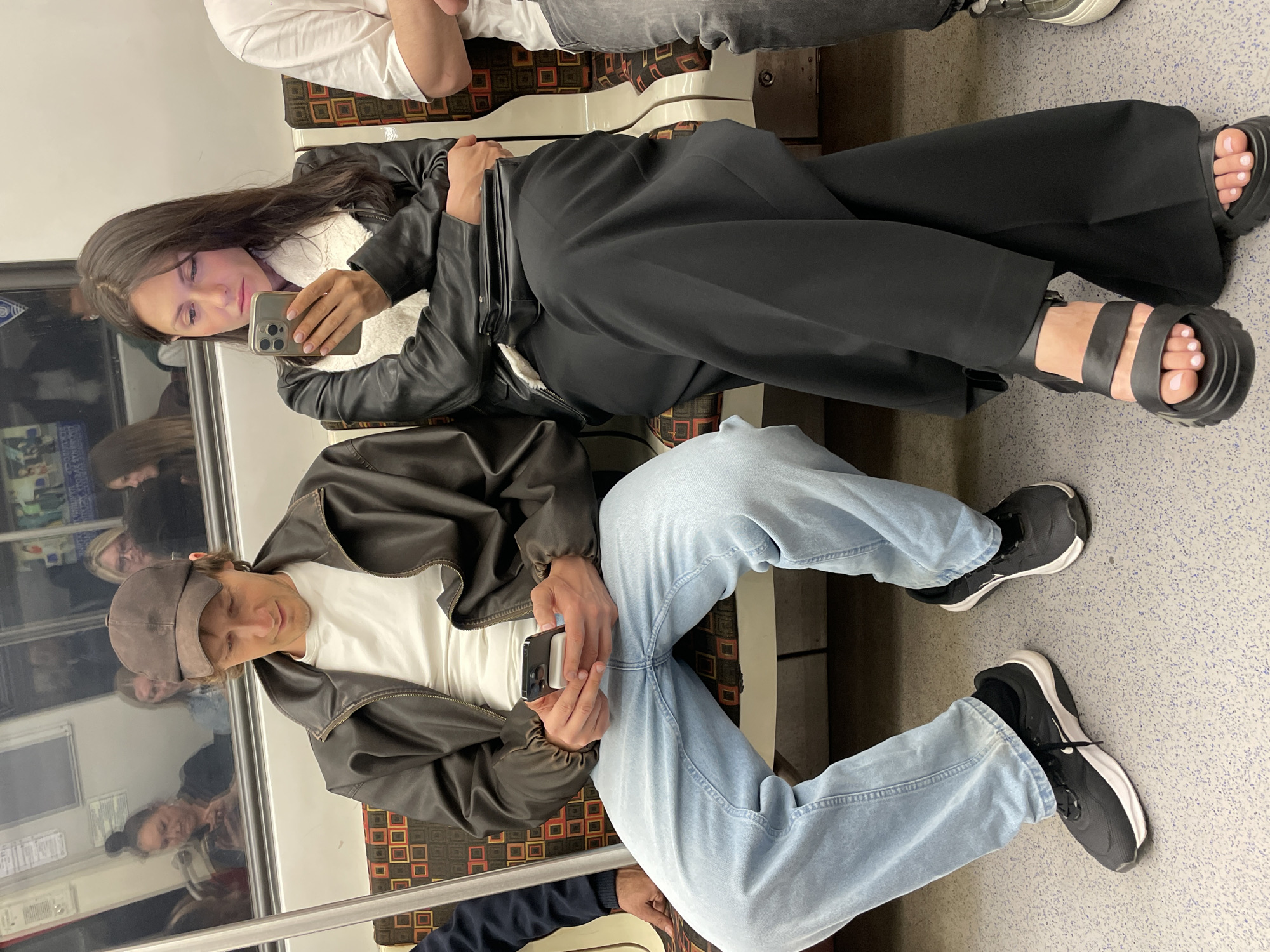 In the city of Unknown, Unknown, two individuals are seated on a subway train, both engrossed in their mobile phones. On the left, a man wearing a brown baseball cap, a brown leather jacket over a white t-shirt, and light blue jeans, holds a black smartphone in his hands. To his right, a woman with dark hair, dressed in a black leather jacket with a white fuzzy collar and black wide-leg trousers, also looks at her phone. The seating area of the train features a patterned upholstery with squares of orange, brown, and cream. The environment appears to be a subway car, indicated by the reflective surface behind the passengers showing other commuters and the train's interior.