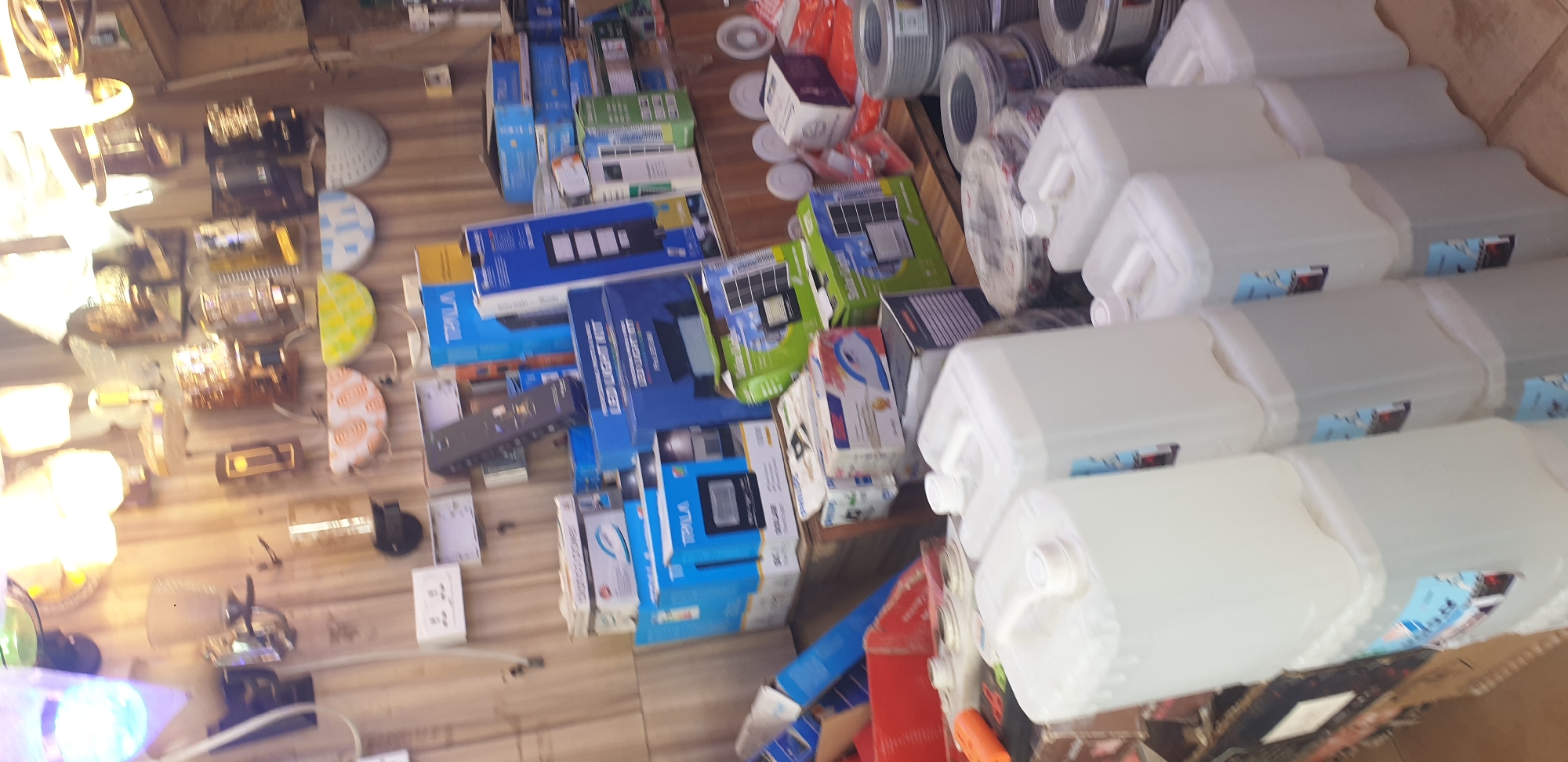 The image captures an indoor commercial or storage space, likely a shop specializing in electrical supplies and household goods, located in Sabon Tasha, Nigeria. The photograph itself appears to be rotated 90 degrees counter-clockwise.
The scene is densely packed with a wide array of products. On the left side, numerous lighting fixtures are displayed, including various decorative lamps, chandeliers, and possibly wall-mounted lights. Among these are distinct half-moon shaped decorative pieces, one with a lime green pattern and another with an orange design. Electrical components such as white power outlets, switches, and a small black remote control are also visible on what appears to be a light-colored wooden surface.
The central and right-central areas are filled with stacks of packaged goods. Prominent among these are multiple blue cardboard boxes, many bearing the brand name "TRILA" and one clearly labeled "LED LIGHT KIT". Other boxes, in green and blue packaging, contain what appear to be solar panel systems, with partial text indicating "SOLAR" and possibly "Philips".
On the far right, a large stack of identical, rectangular, white plastic containers with handles and screw-top lids is visible. Each container features a blue and white label. Above these, several large spools of electrical wire or cable are present.
No people are depicted, suggesting a focus on inventory or product display. The bright lighting indicates the scene is either well-lit by artificial lights or captured during daytime. The overall impression is that of a bustling commercial environment showcasing a diverse range of lighting and electrical products.