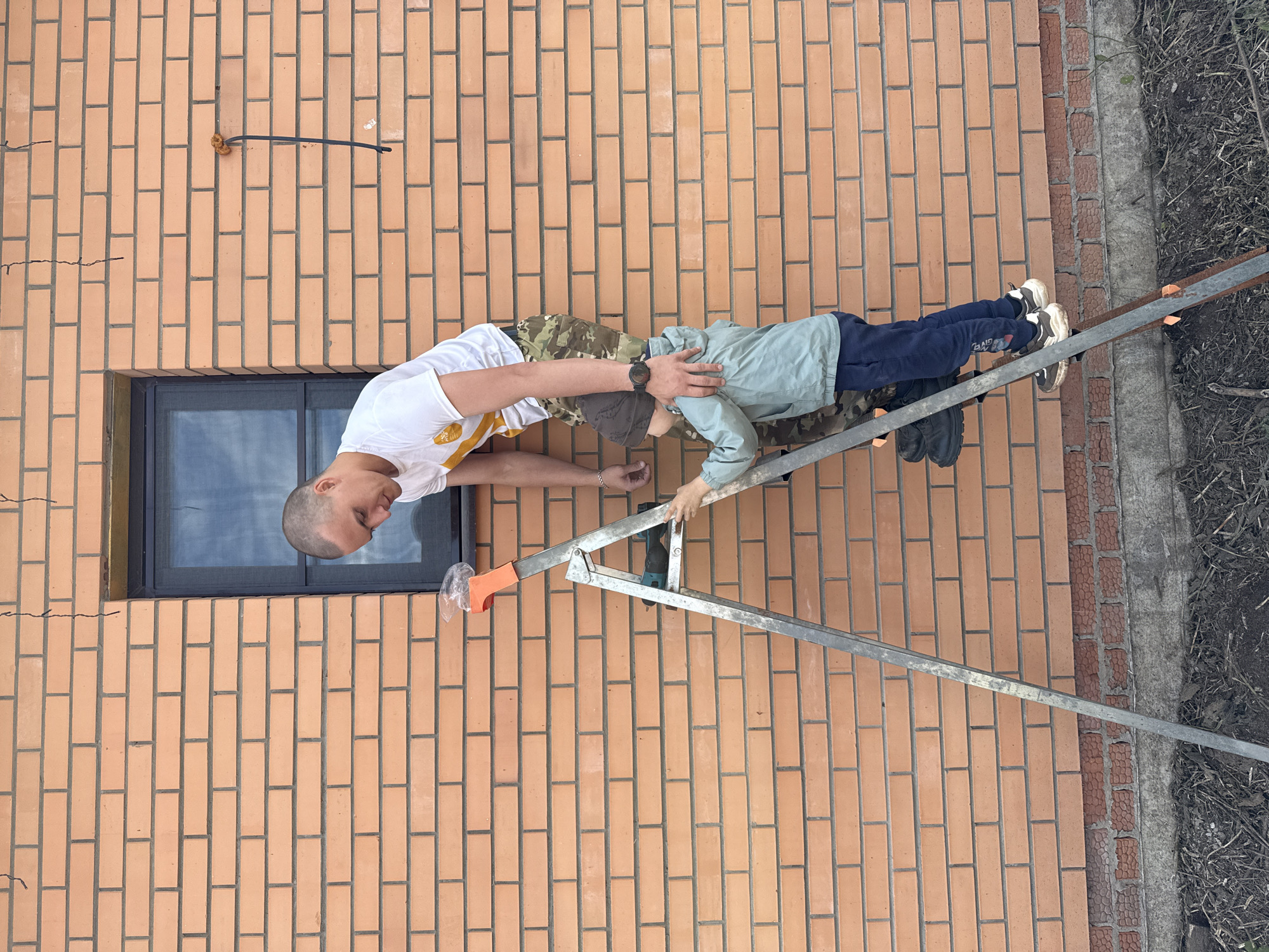 An overhead outdoor shot depicts a man and a boy on a ladder against a brick wall. The man, positioned on the left, wears a white t-shirt, camouflage pants, and a watch. He is holding the boy’s shoulder. The boy, wearing a light blue shirt and dark blue pants, is further up the ladder, reaching towards the wall. An aluminum ladder with orange caps rests against the brick wall. The wall has a dark-framed window on the left side. To the right of the wall is a concrete foundation with dirt and dry vegetation. A black wire runs across the brick surface above the window. The image was taken in Chaikovsky, Russia.