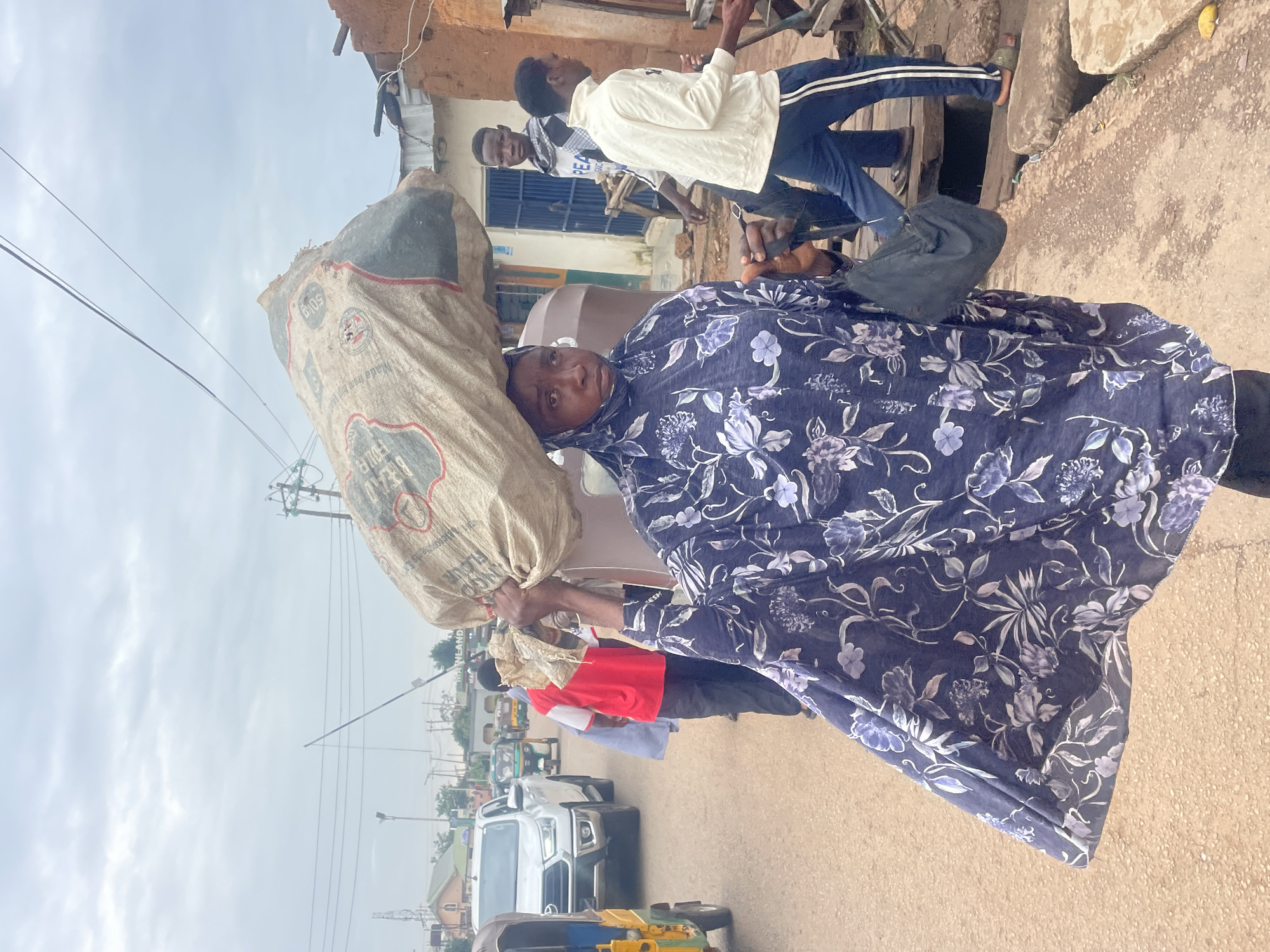 In Gura Top, Nigeria, a woman wearing a blue floral headscarf and dress carries a large sack of grain on her head. She is walking on a dirt road, past a line of vehicles and a few other people. The sky is overcast, and the overall scene appears to be a typical day in a Nigerian city.