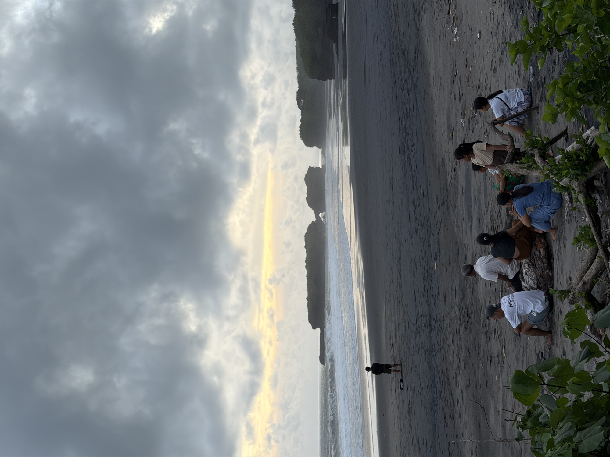 The image depicts a beach scene in Selemadeg Timur, Indonesia. A group of six individuals are located on the right side of the frame near some vegetation. They appear to be engaged in some activity on the sand, bending over with their hands near the ground. Further down the beach, closer to the water, is a lone figure standing near the shoreline. The beach itself is dark sand, possibly volcanic. The water is a mix of light and dark tones, suggesting varying depths or wave patterns. In the distance, there are rock formations jutting out of the water, topped with greenery. The sky is predominantly cloudy, with a gradient from dark gray on the left to a lighter, possibly sunlit area near the horizon.